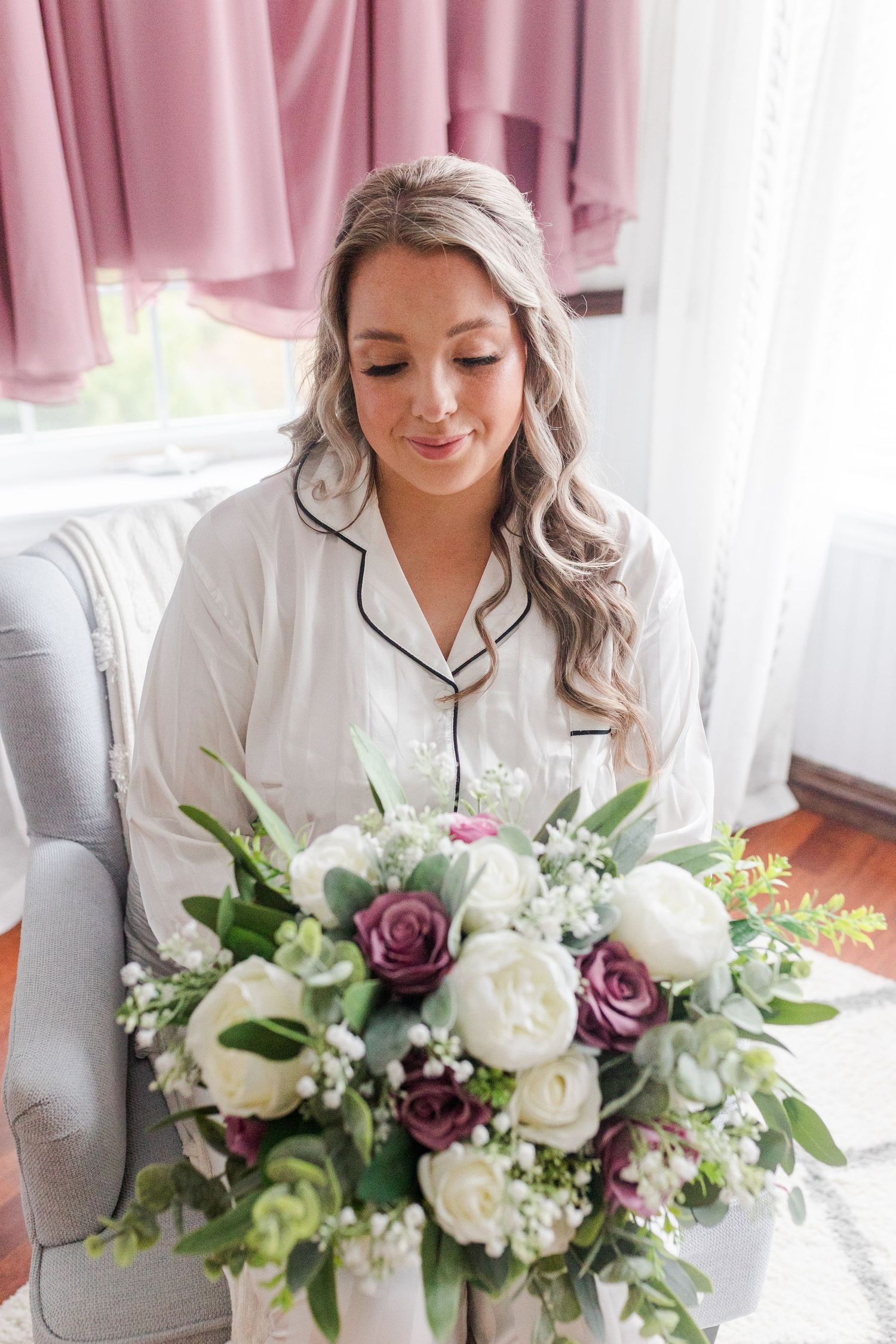 Woman in white silk robe holds beautiful wedding bouquet with white and purple flowers indoors by window.