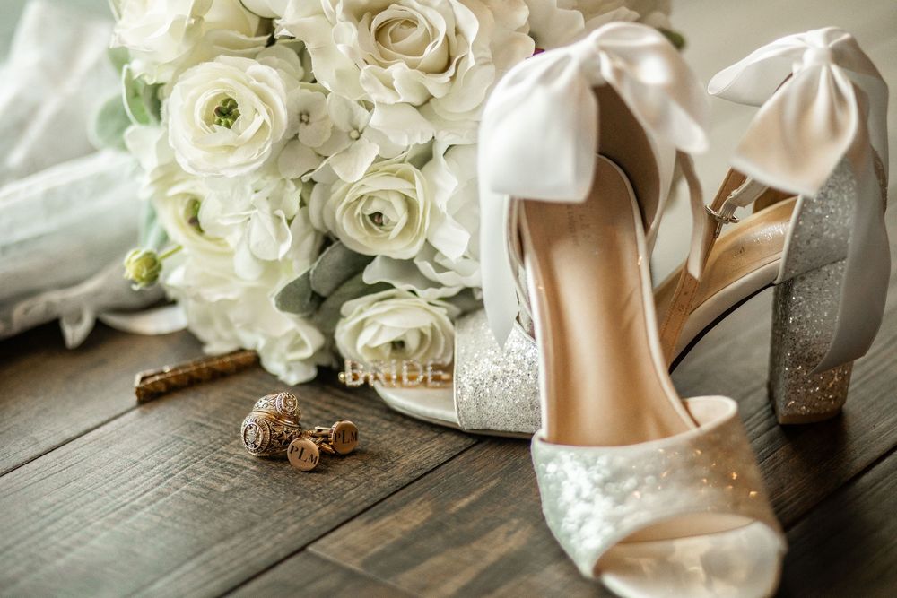 Wedding details featuring sparkling white heels next to cream roses and acorns on rustic wooden floor.