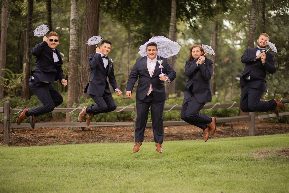 Groomsmen perform synchronized jumping poses on a grassy lawn during wedding party photos.