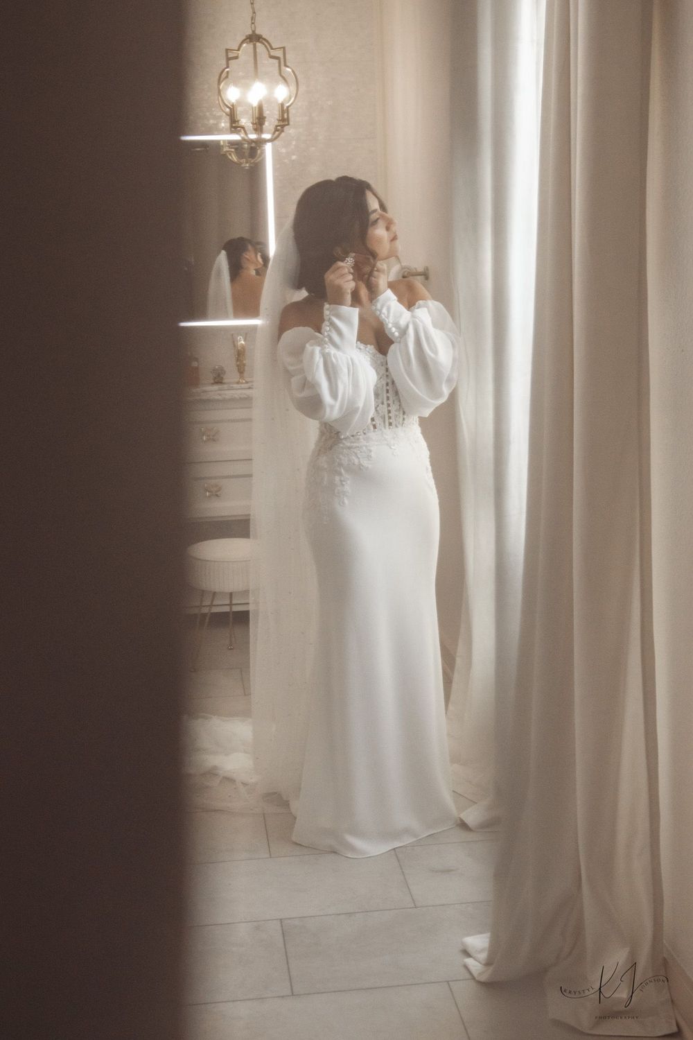 A bride in a flowing white dress poses elegantly against light-filled curtains in an indoor setting.