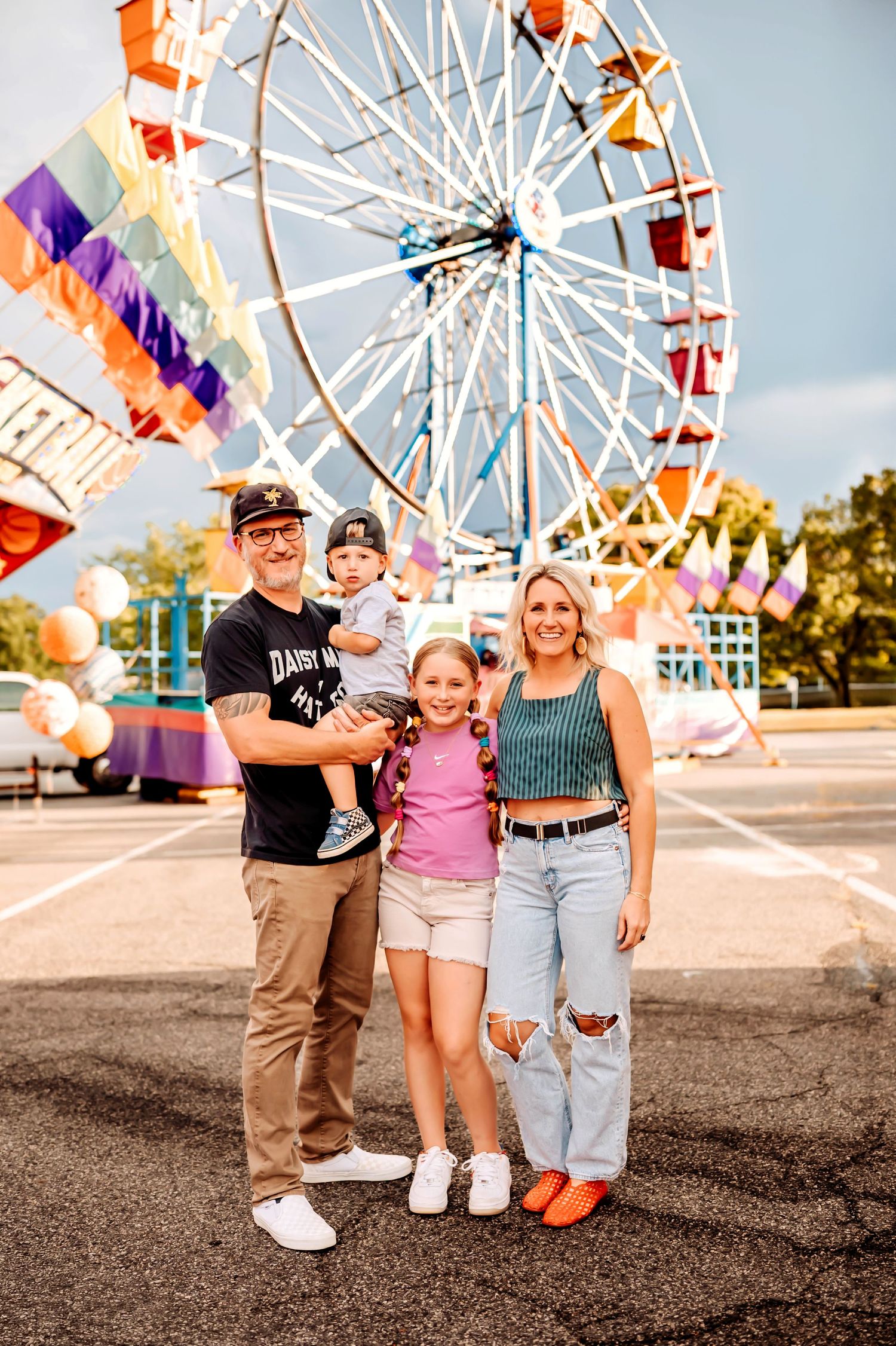 A family enjoys a summer day at a carnival with a colorful ferris wheel in the background.