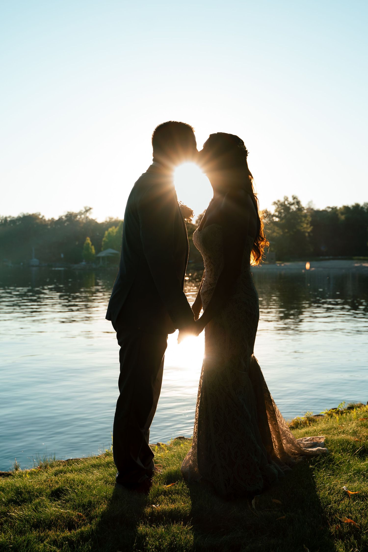 Silhouetted couple standing by a lake at sunset creating a romantic backlit series of portraits against the water.