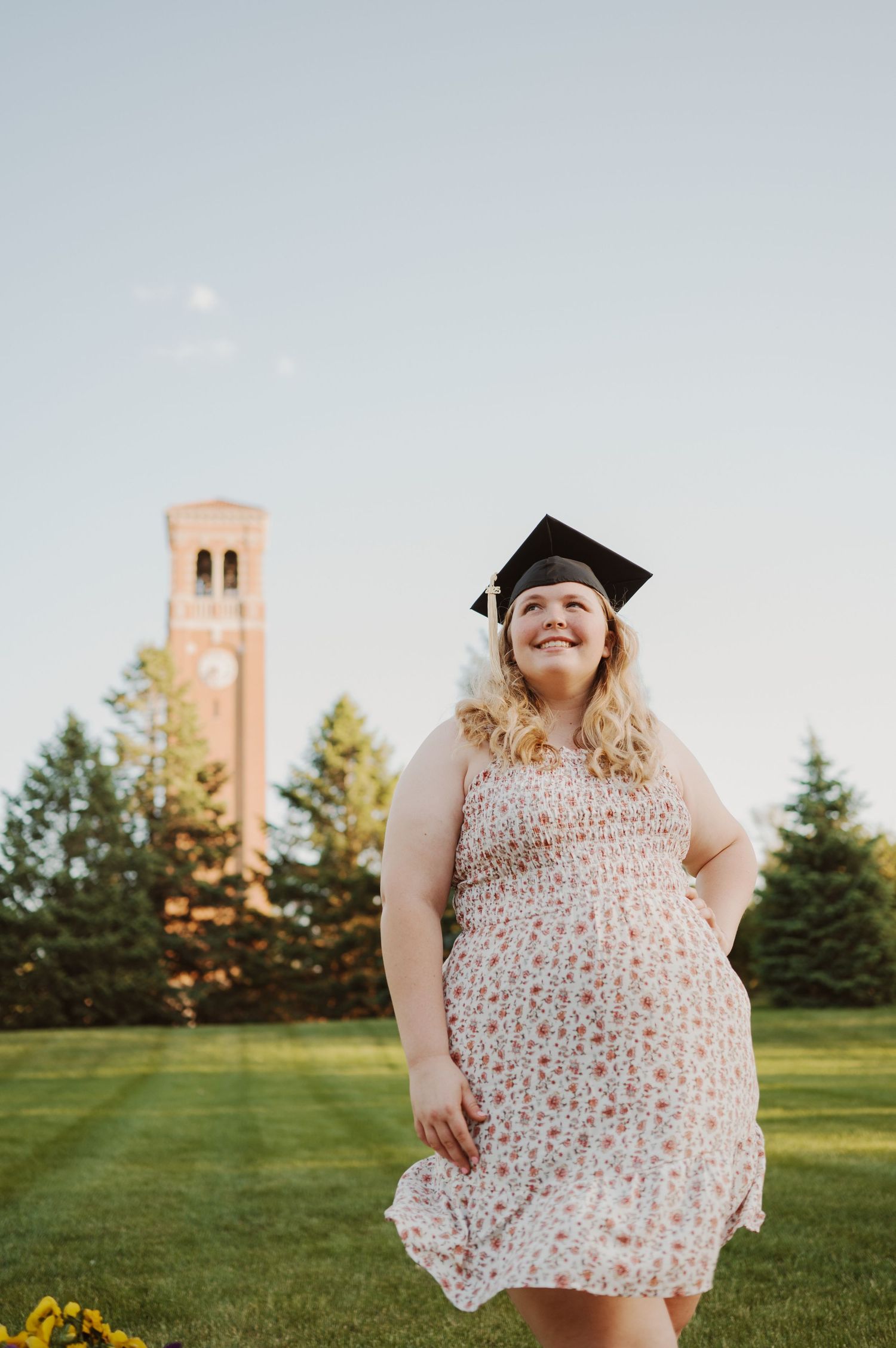 Graduate posing in a white floral dress on a sunny lawn with a brick bell tower in the background.
