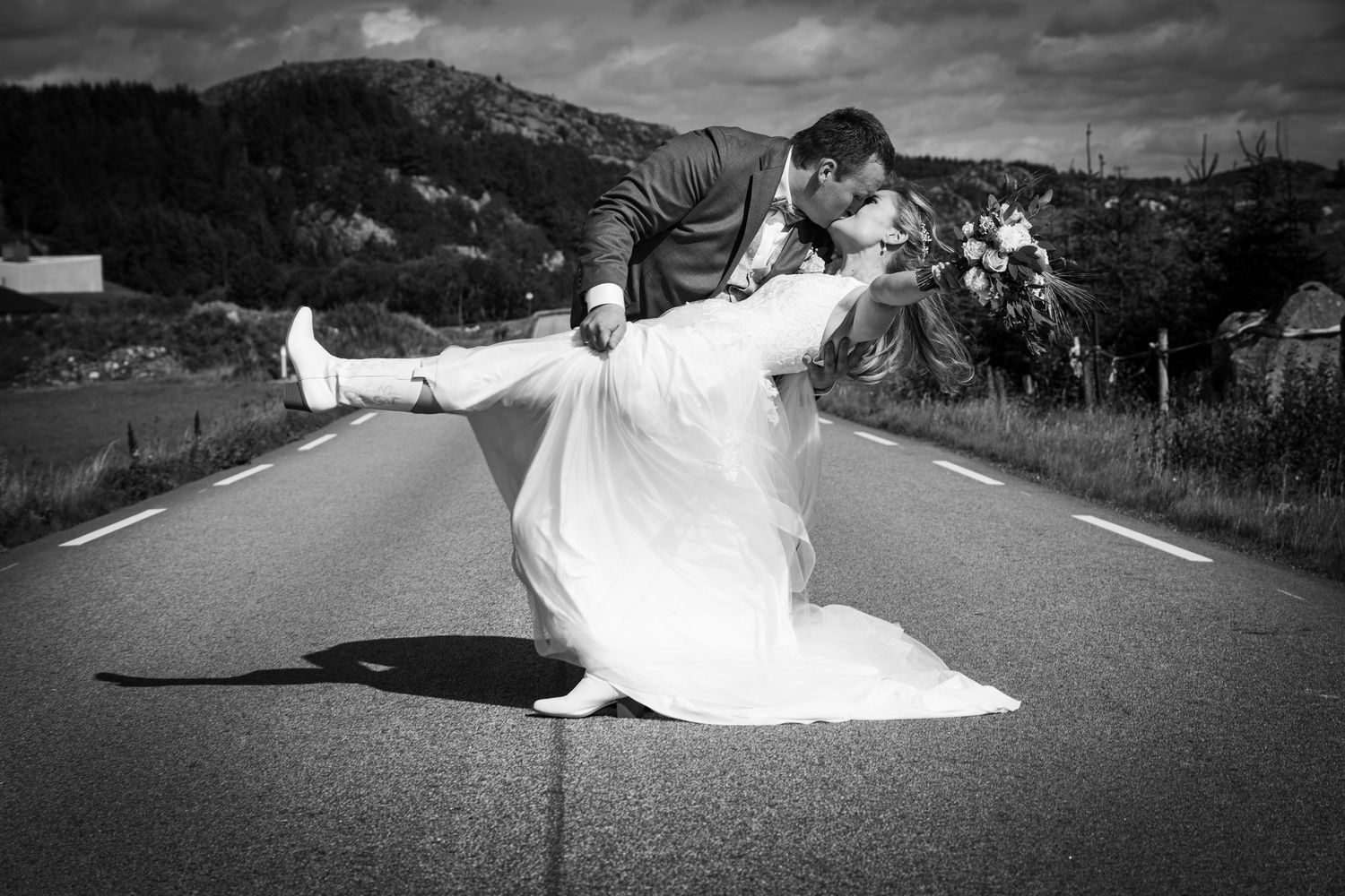 Bride and groom share dramatic dip kiss on open road in stunning black and white photo.