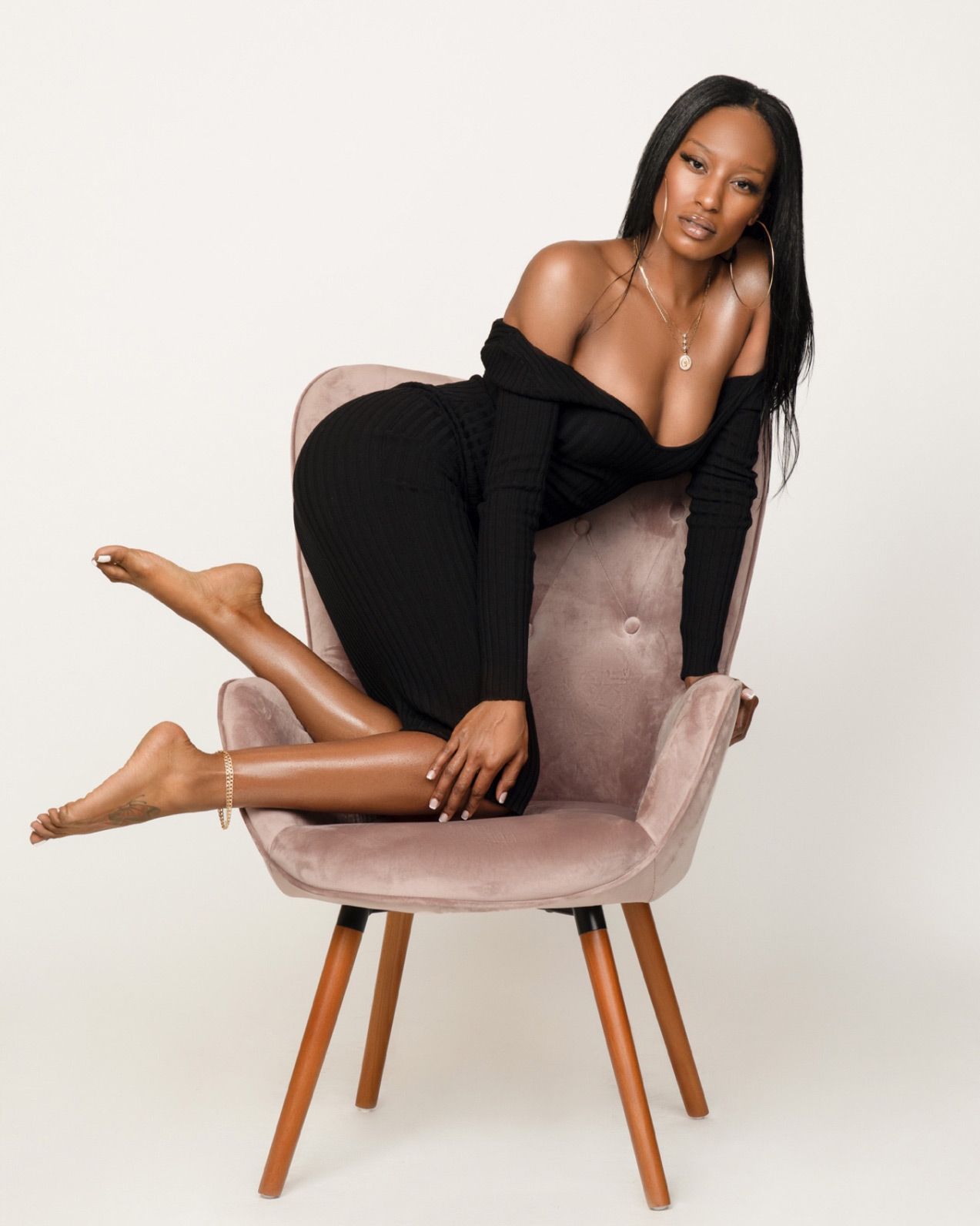 A stylish figure in a black off-shoulder outfit poses elegantly on a pink velvet mid-century modern accent chair.