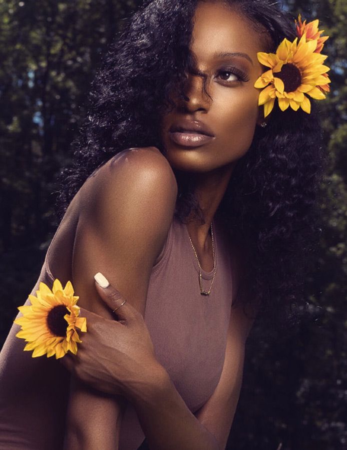 Artistic portrait with sunflowers featuring brown outfit and natural curly hair in dark outdoor setting.