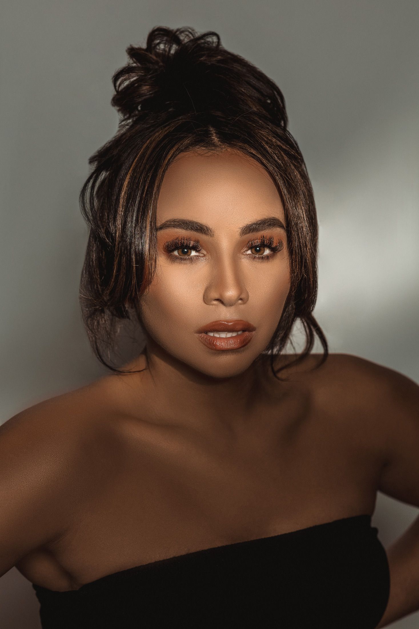 Portrait in dramatic lighting showcasing glowing makeup and a black off-shoulder top against a shadowed background.