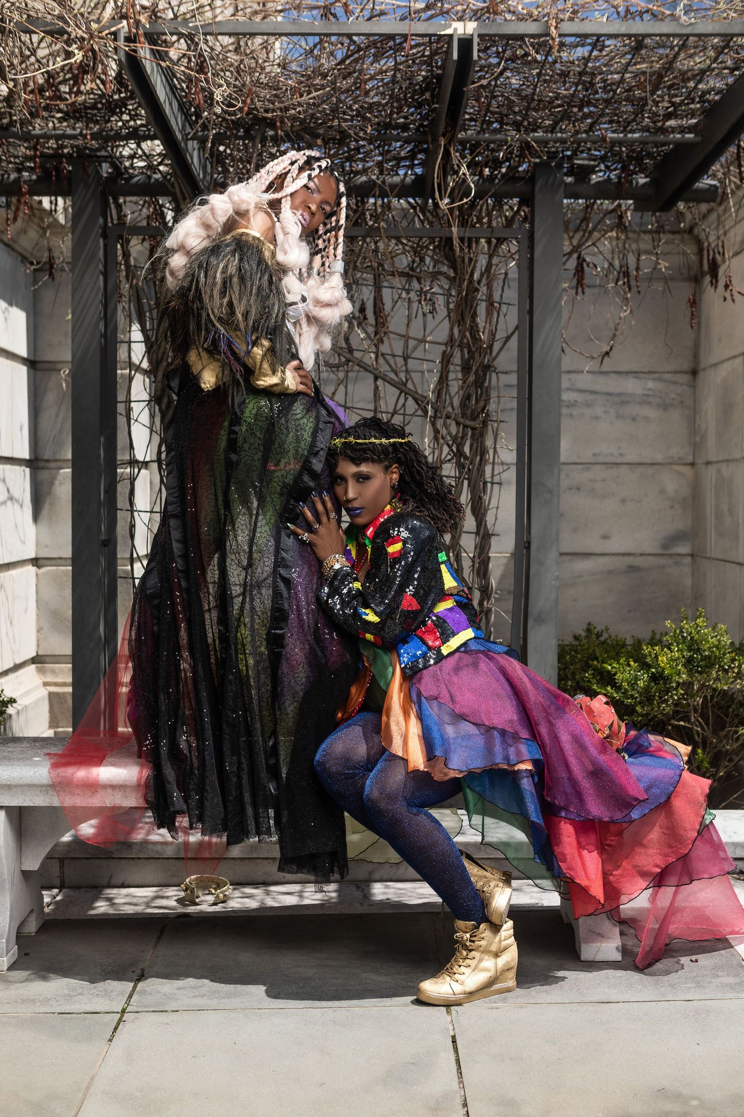 Two models in colorful theatrical costumes pose dramatically under a vine-covered archway during an outdoor photoshoot.