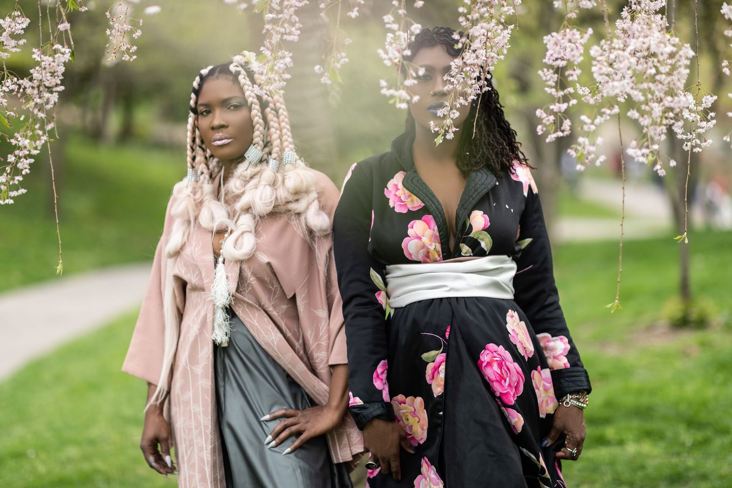Two women in stylish outfits walk together beneath blooming cherry blossoms on a spring day.