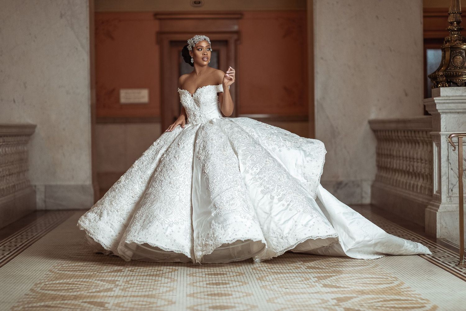 A bride in a flowing white ballgown dress poses elegantly on a marble staircase in a grand architectural setting.