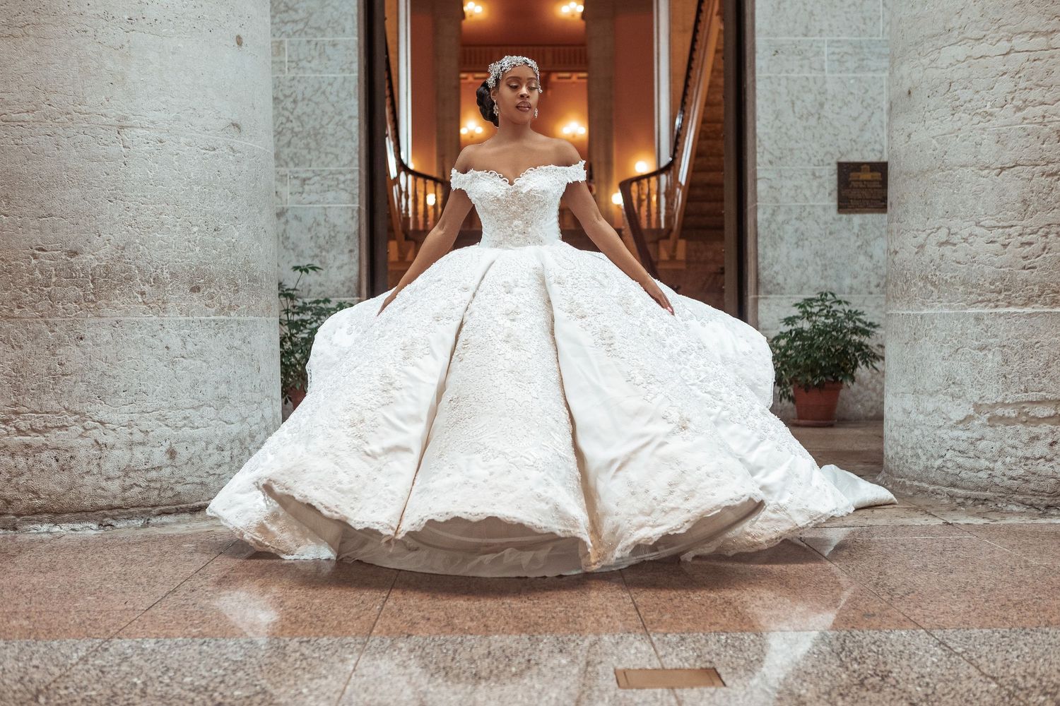 A stunning off-shoulder ballgown wedding dress with intricate lace details billows dramatically on polished stone floor.