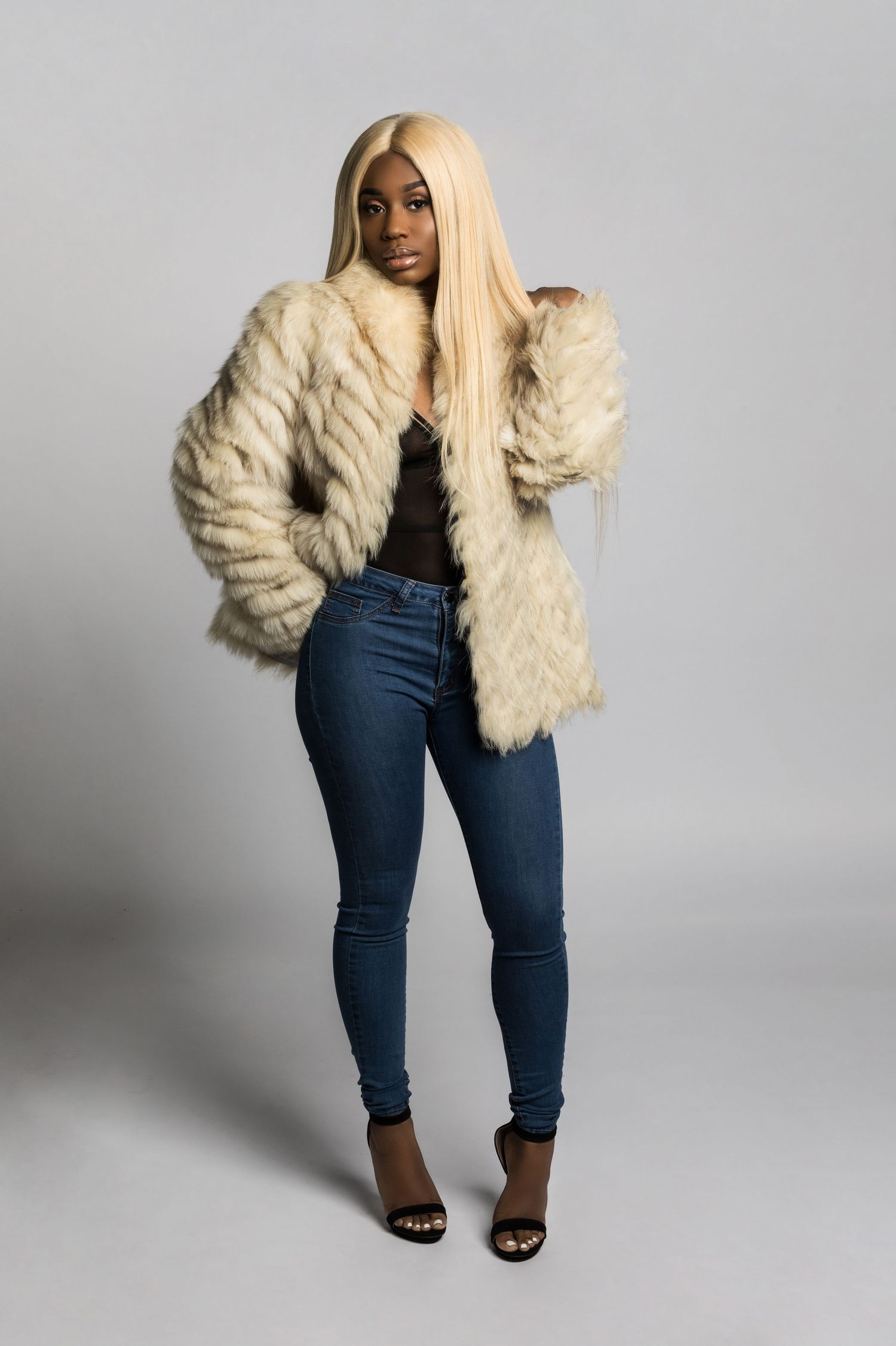 A person wearing a cream fur coat, dark shirt, skinny jeans and heels poses against a gray studio backdrop.