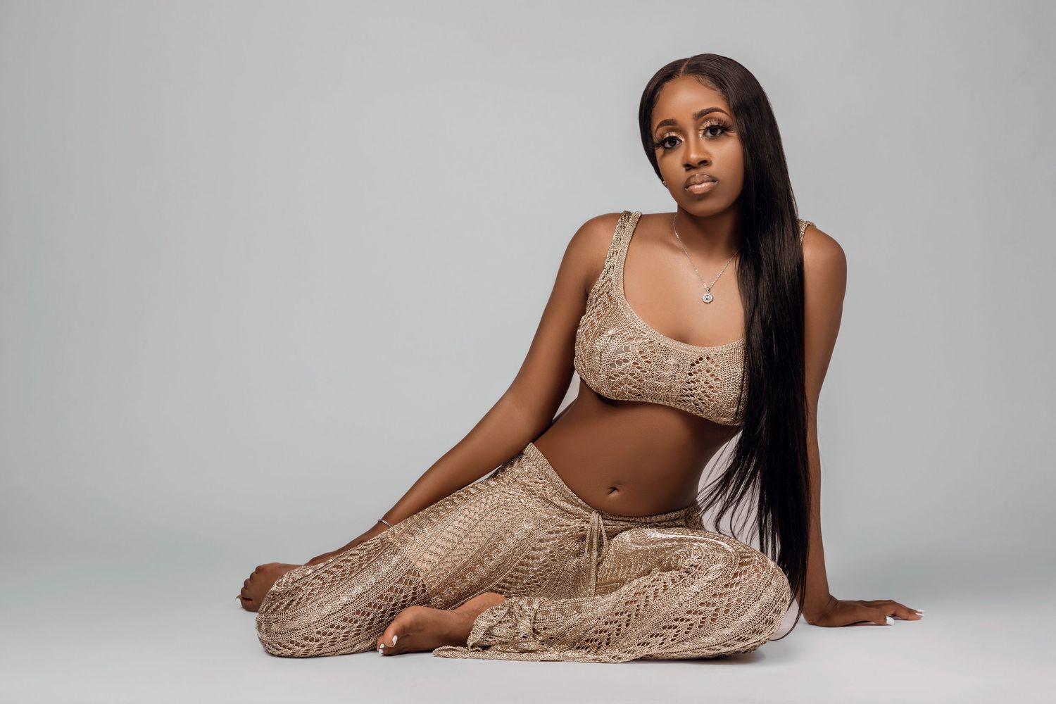 Model in beige knitted two-piece outfit with long straight hair poses sitting on studio floor.