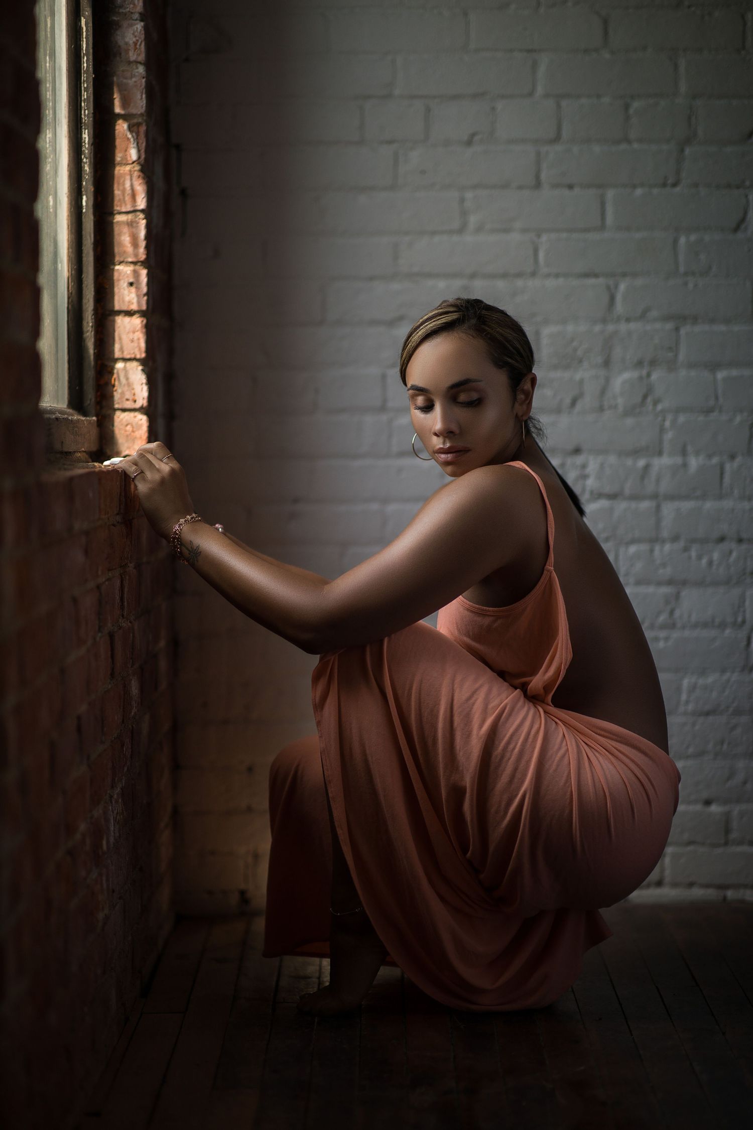 A model in a flowing peach dress crouches by a window in moody lighting against a white brick wall.