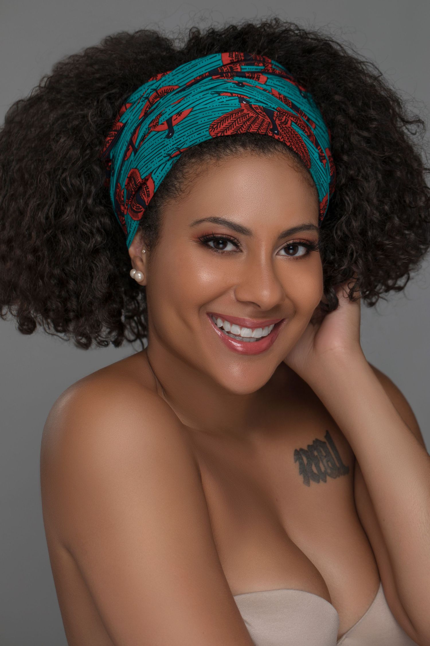 Beautiful portrait featuring natural curly hair, turquoise patterned headwrap, and warm smile against grey backdrop.