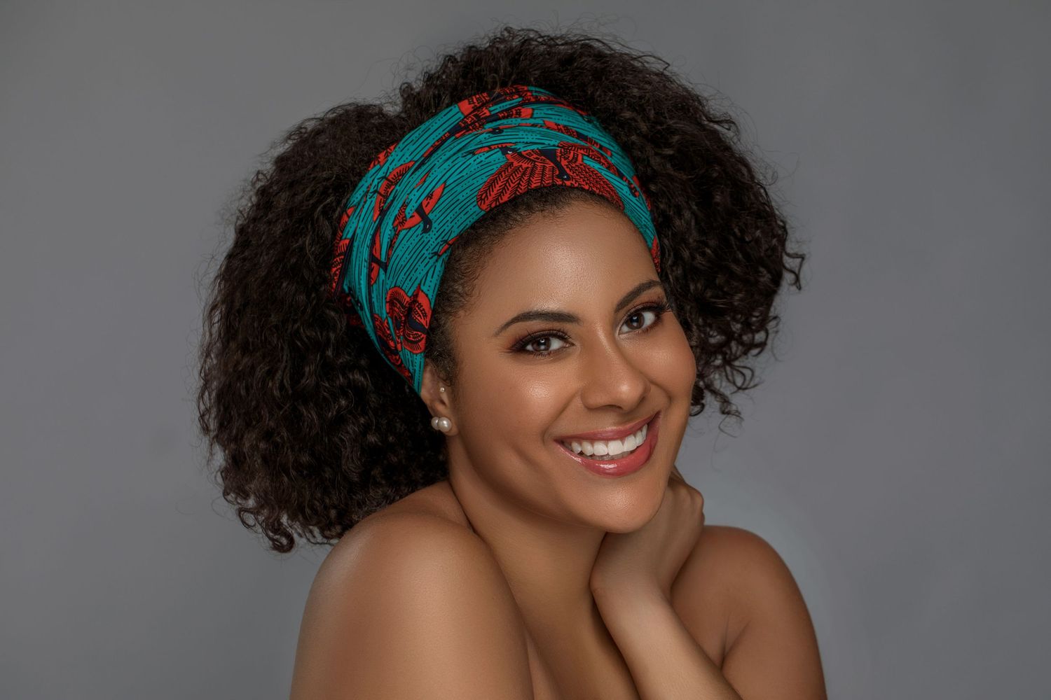 Person with natural curly hair wearing a turquoise and coral patterned headband smiles against grey backdrop.