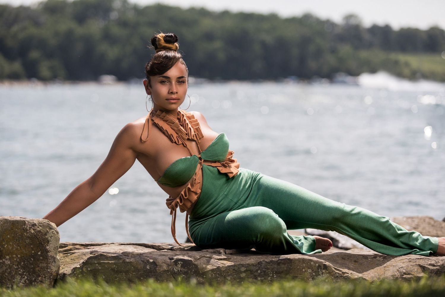 Athletic model in emerald green workout attire poses on rocky shore with lake and trees in background.