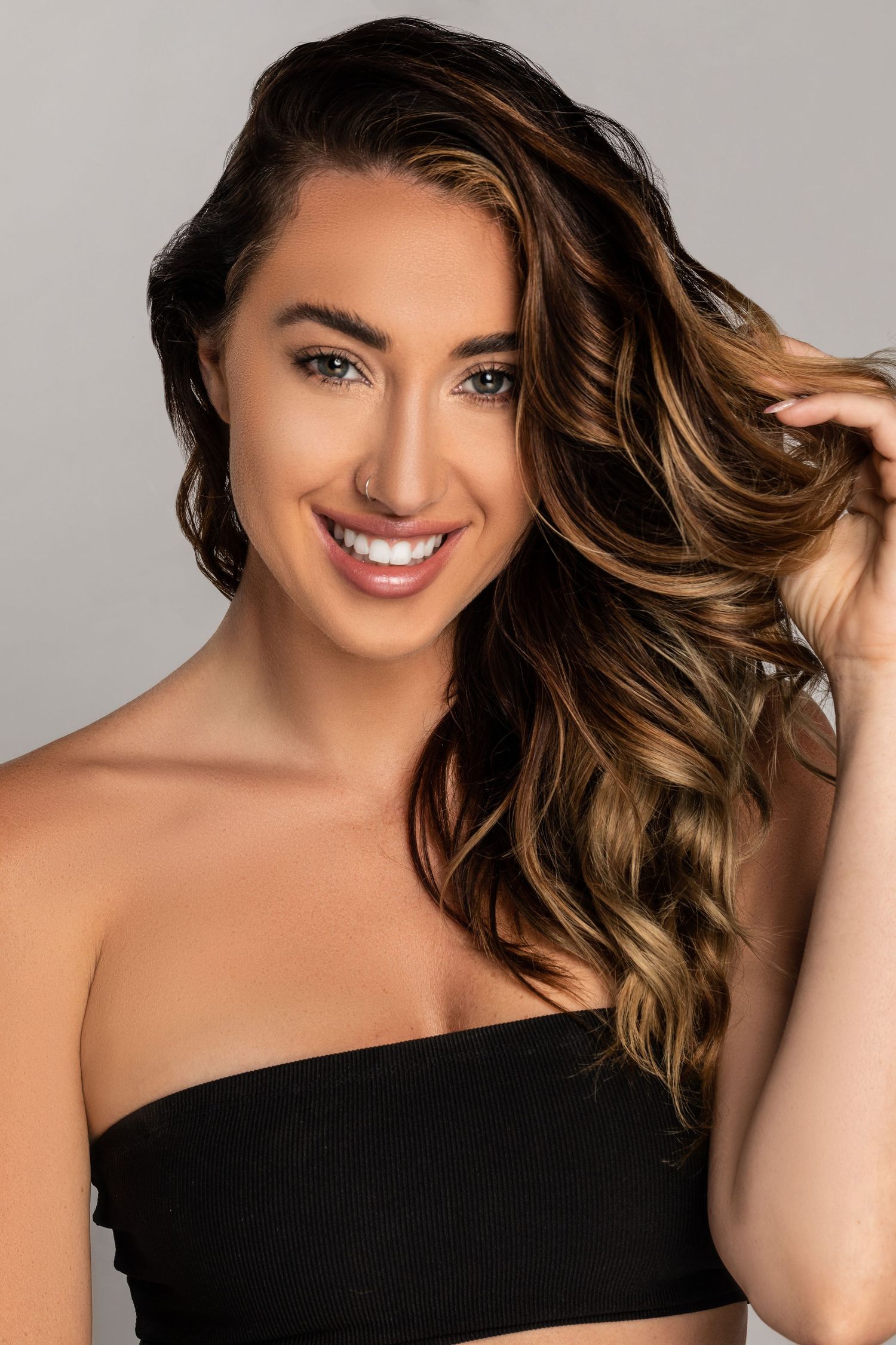 Beauty portrait with flowing caramel highlighted hair wearing black top against neutral background.