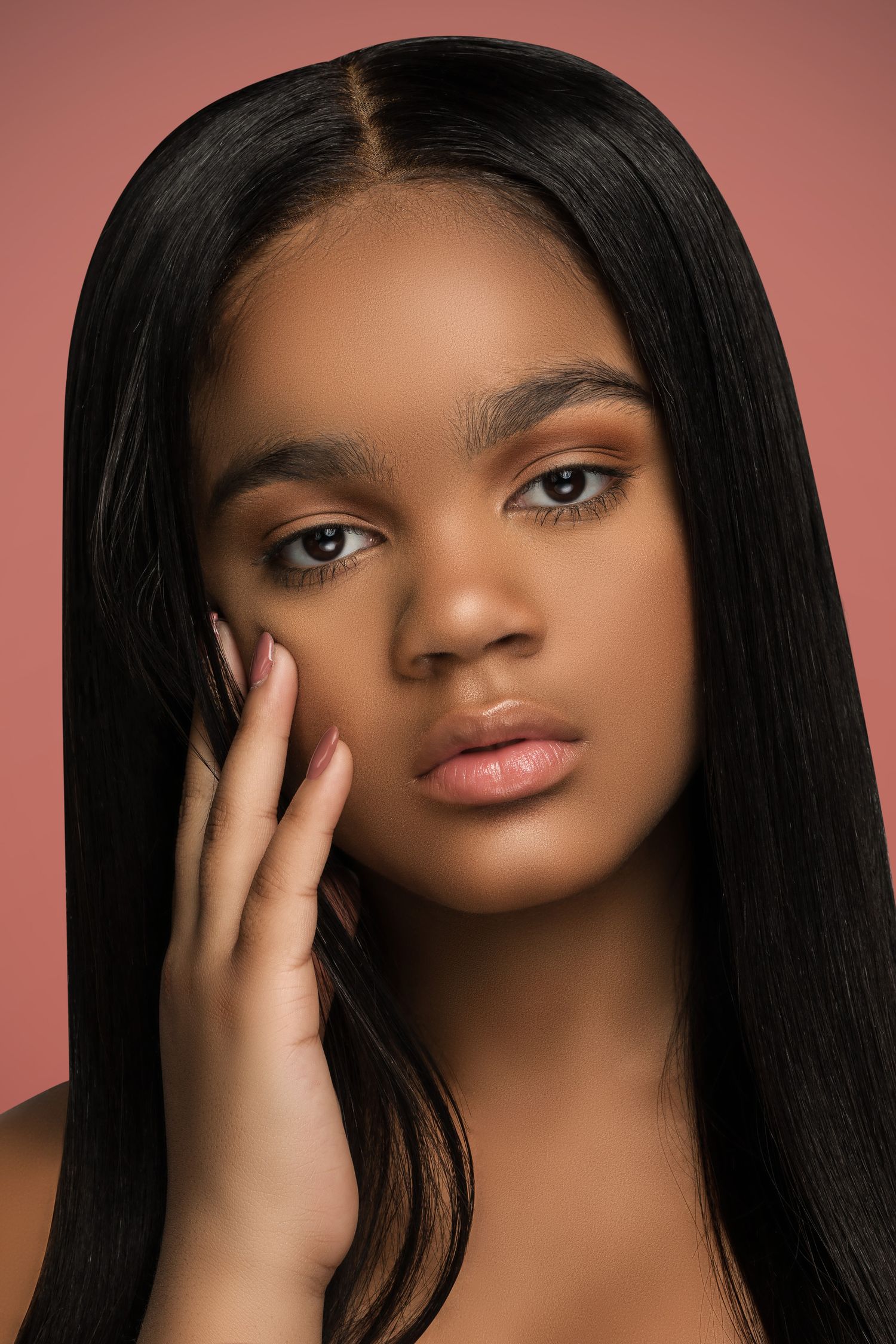 Beauty portrait with flawless glowing skin, long straight dark hair, and soft neutral makeup against warm brown background.