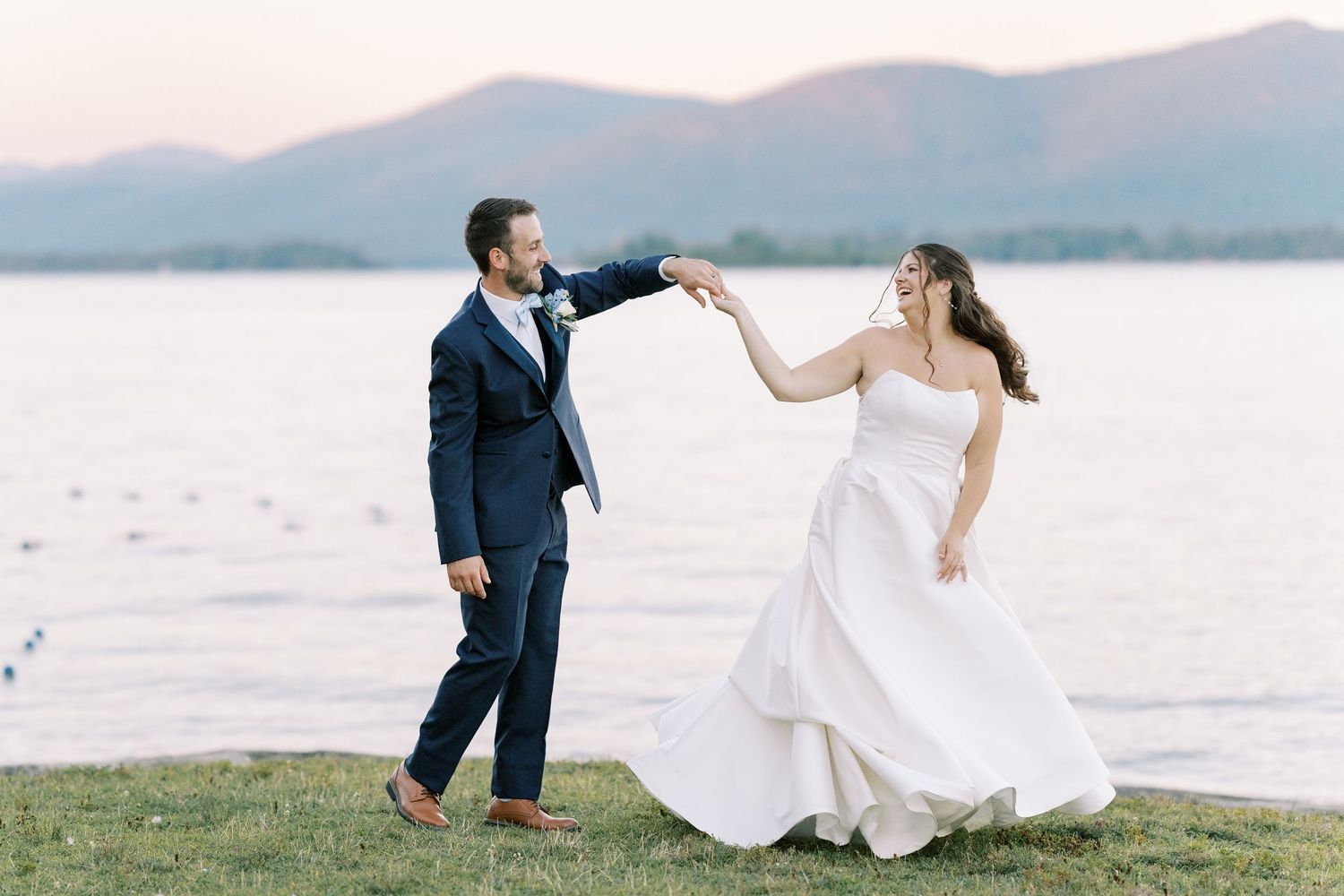 A couple in wedding attire dances together on a sandy salt flat with mountains in the background at sunset.