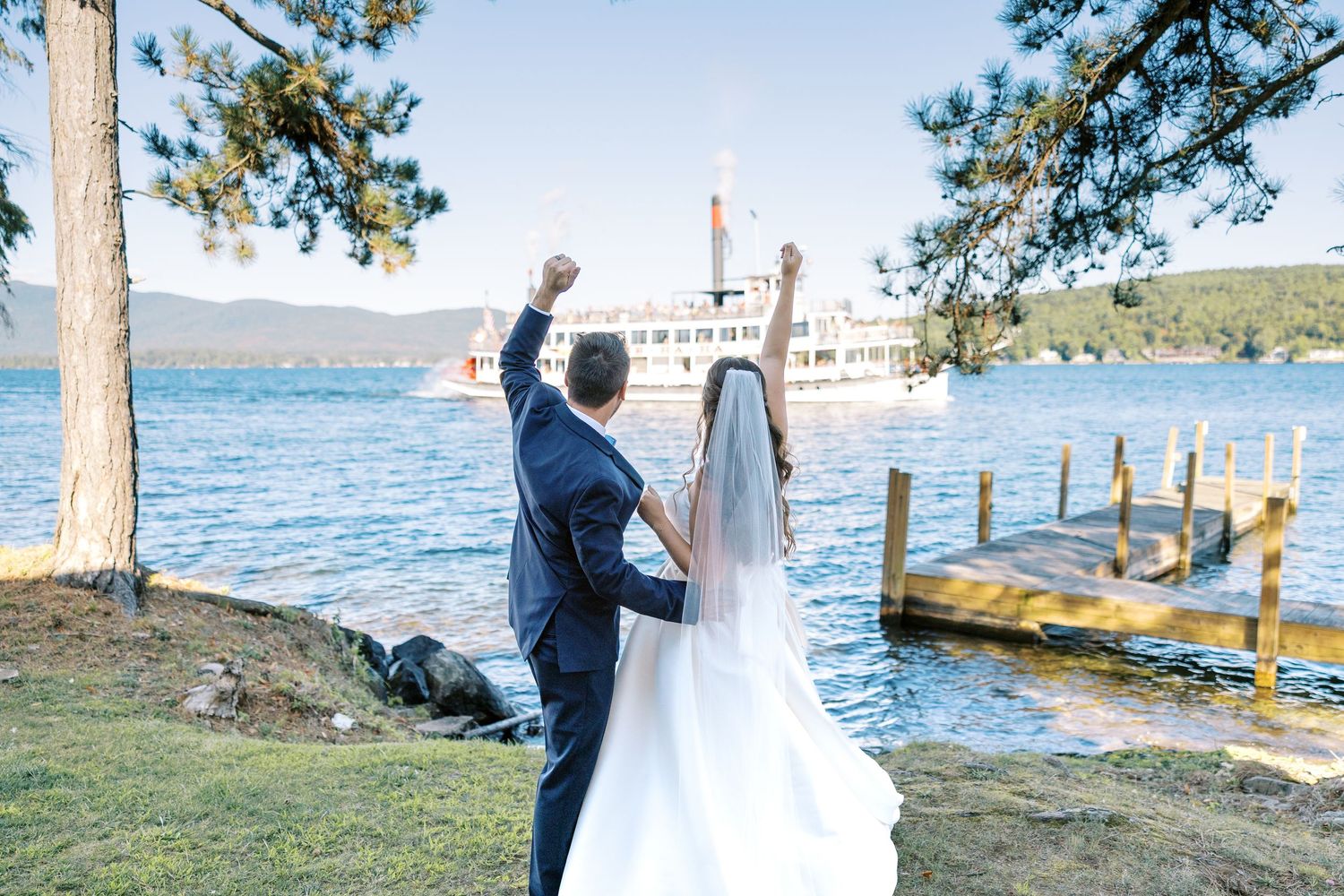 Newlyweds share romantic moments along the scenic lakeside with mountains and pine trees in the background.