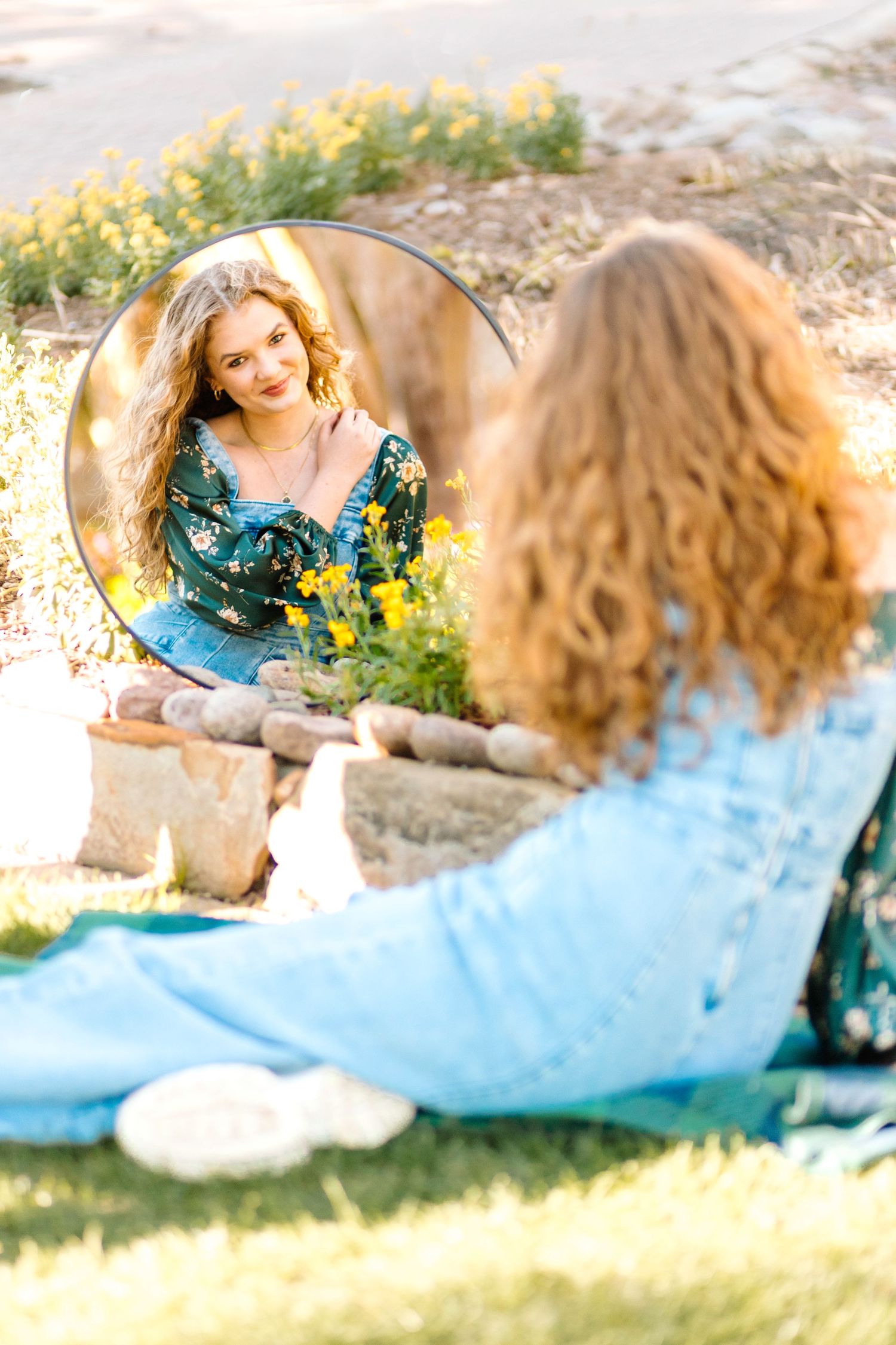 Artistic mirror reflection in nature shows a woman in denim overalls and floral cardigan with sunlit trees in background.