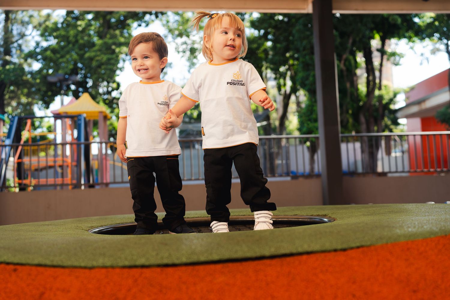 Duas crianças pequenas em pé, juntas, em um playground, vestindo roupas brancas iguais. Campanha para instituição de ensino.