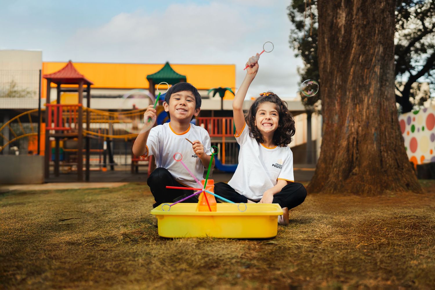 Crianças brincando entusiasticamente com uma caixa sensorial ao ar livre em um playground. Campanha para instituição de ensino.
