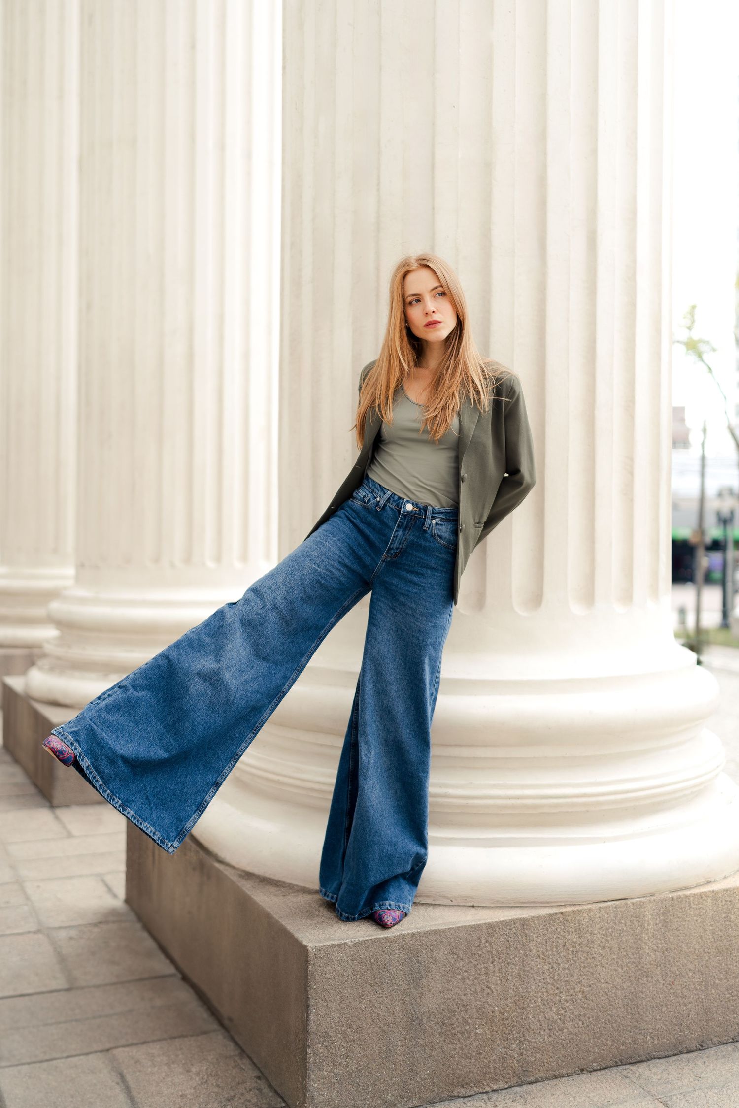 Model in grey jacket and wide-leg jeans poses against white architectural columns.