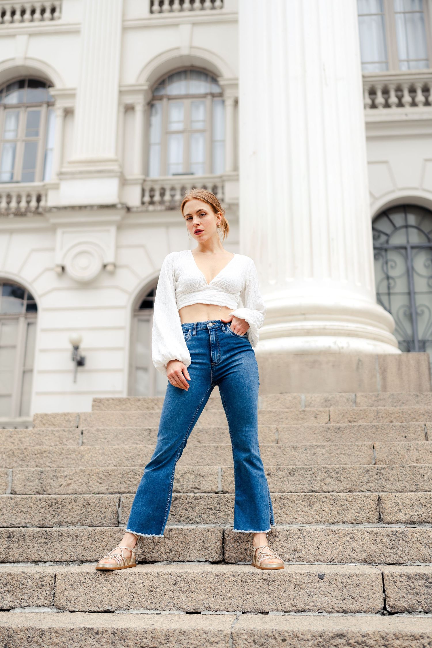 Fashion model poses on stone steps wearing cropped white top, flared jeans and sneakers against classical architecture.