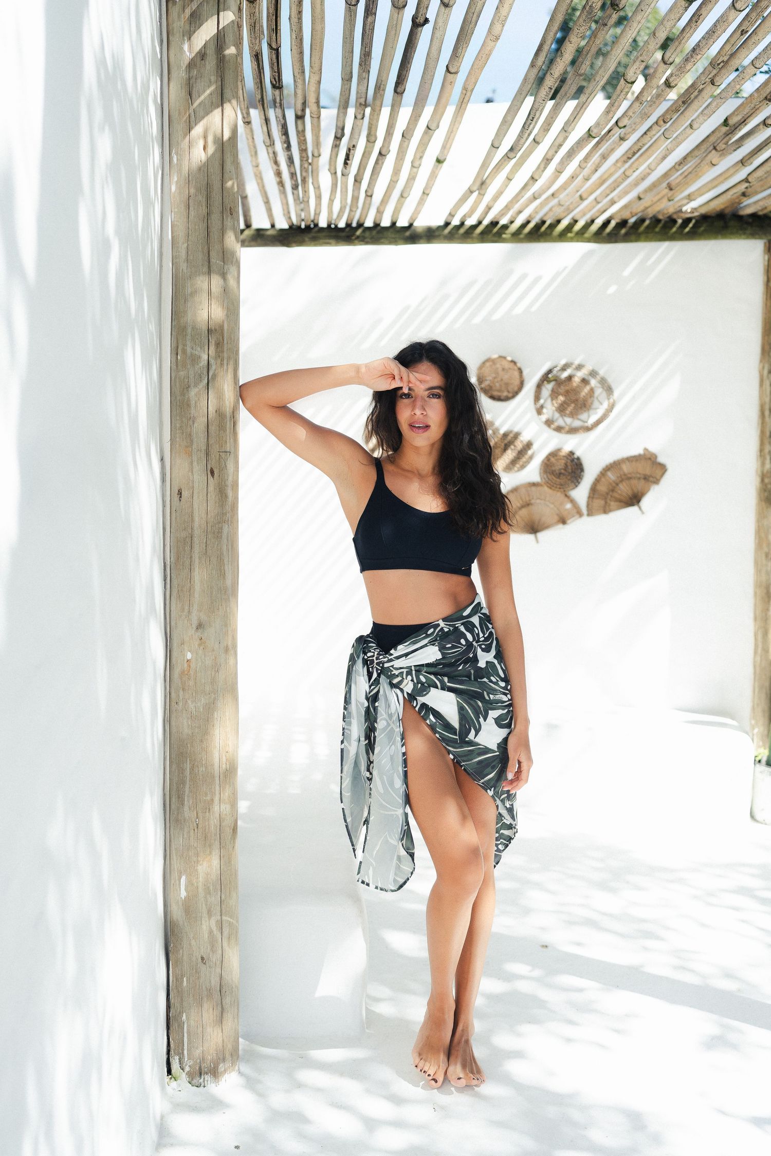 Woman in black crop top and printed sarong poses under wooden pergola with shadow patterns.