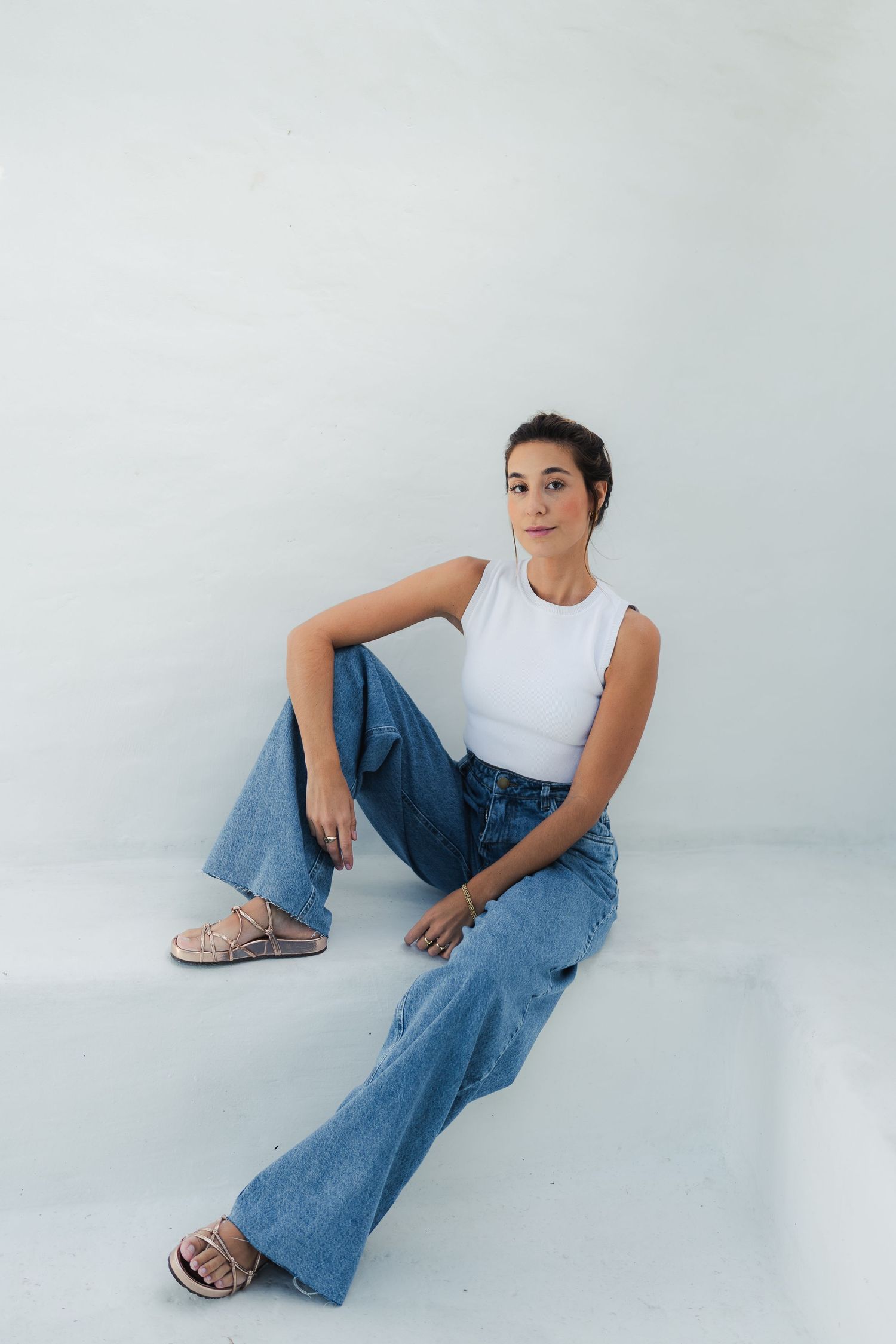 Model in casual white top and jeans sits on white floor.