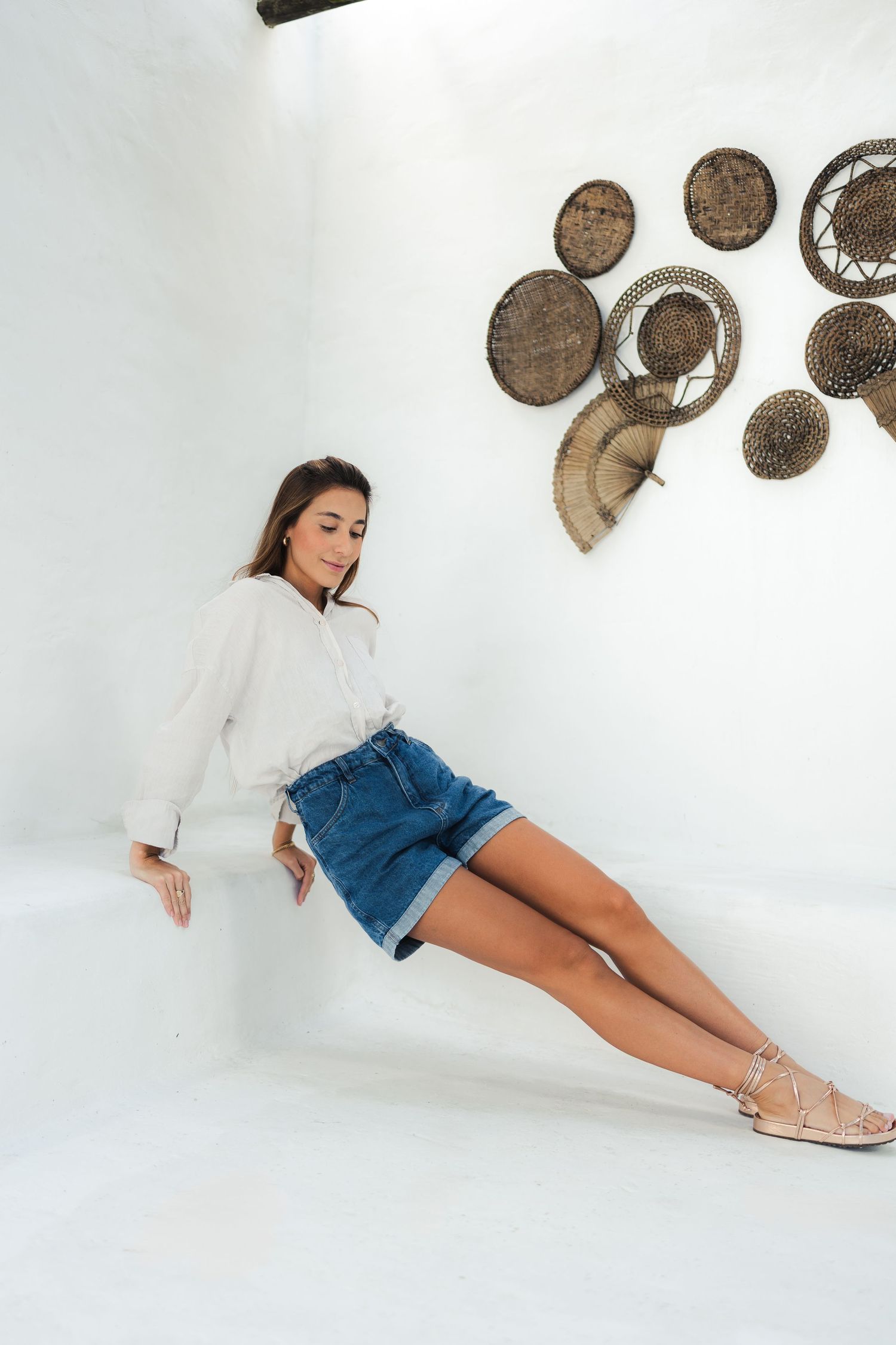Model reclines in denim shorts and white top against white wall with decorative woven baskets.