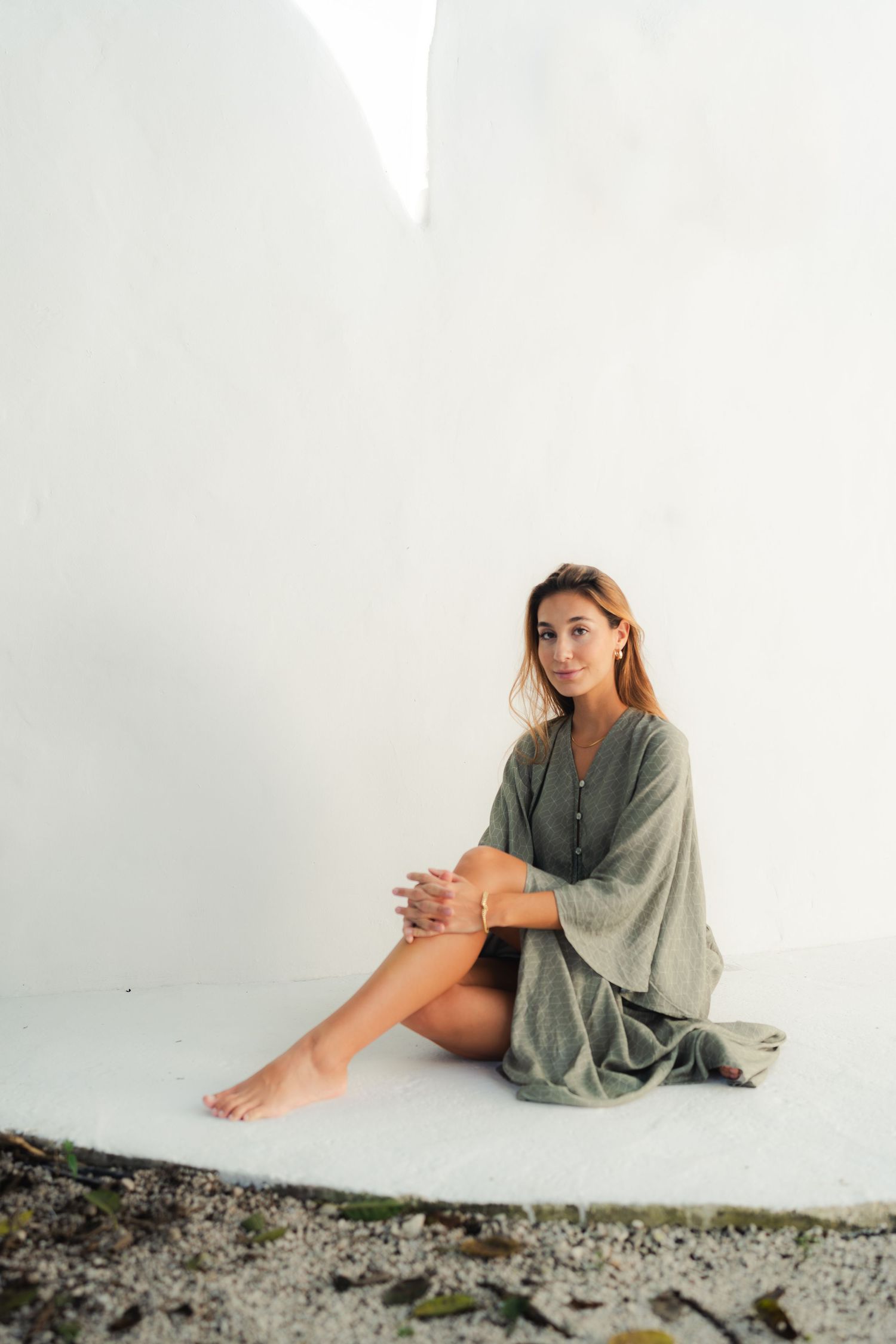 Model in loose gray dress sits casually on ground against white wall.