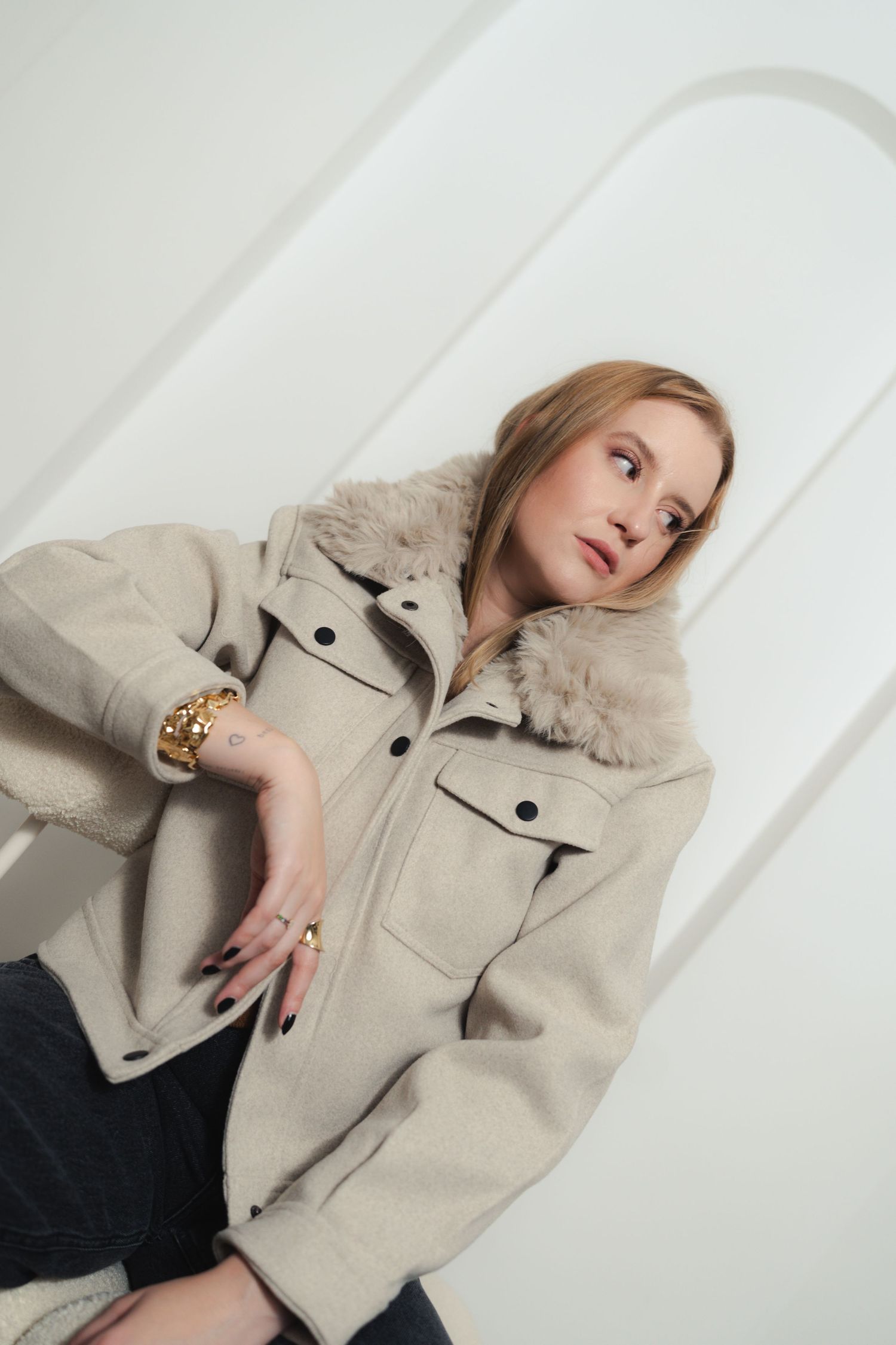 Model wearing beige shearling jacket with fur collar poses against white architectural background.