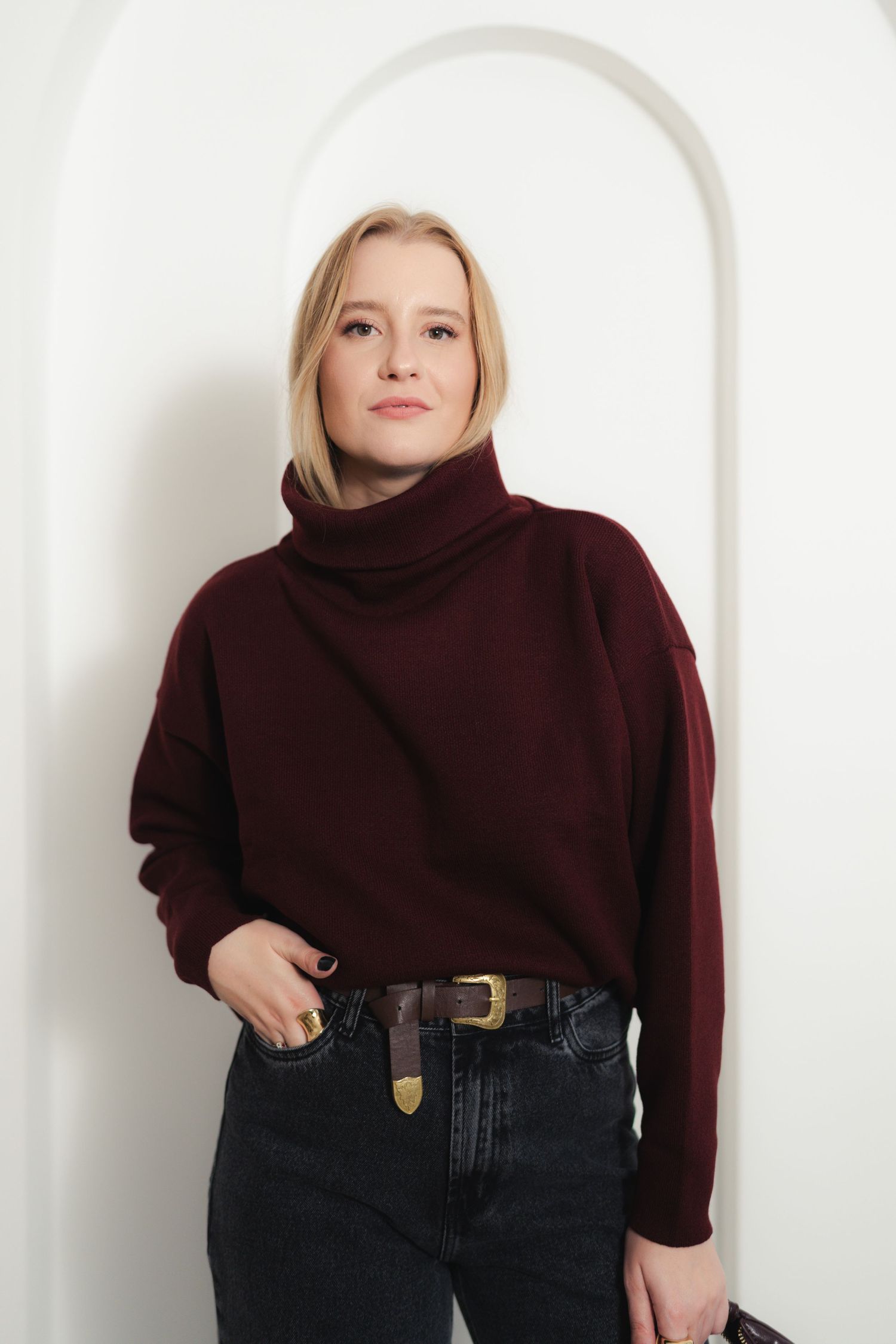 Model wearing burgundy turtleneck sweater with dark jeans and leather belt in minimalist setting.