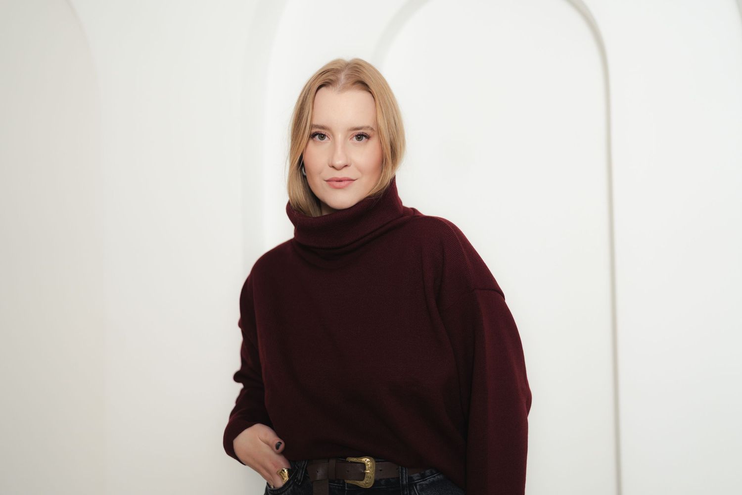 Model poses in burgundy turtleneck sweater with dark jeans against white arched background.