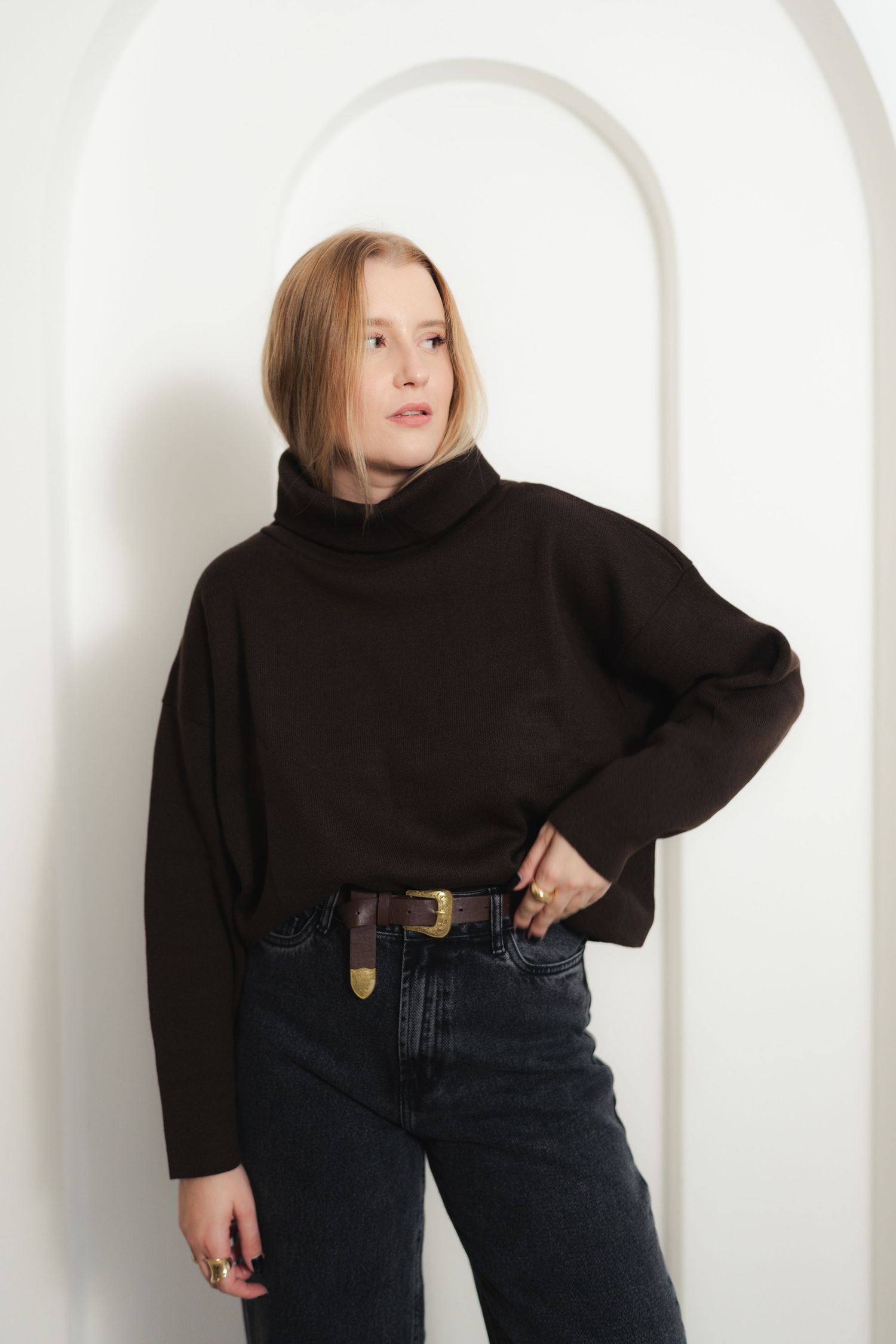 Model in dark brown turtleneck sweater and high-waisted jeans against white wall with arch.