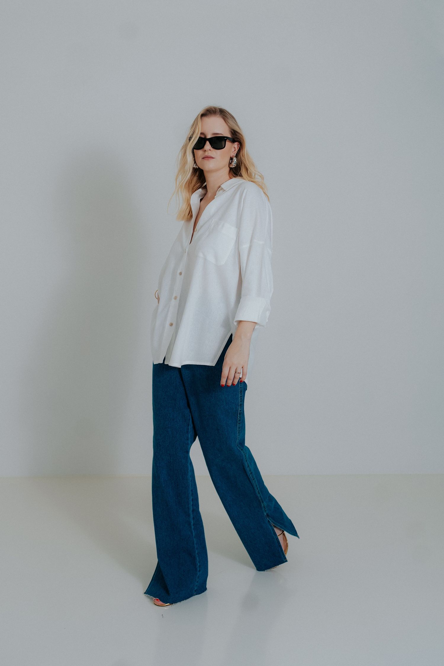 Fashion model wearing white button-down shirt, flared blue jeans and sunglasses poses against light gray background.