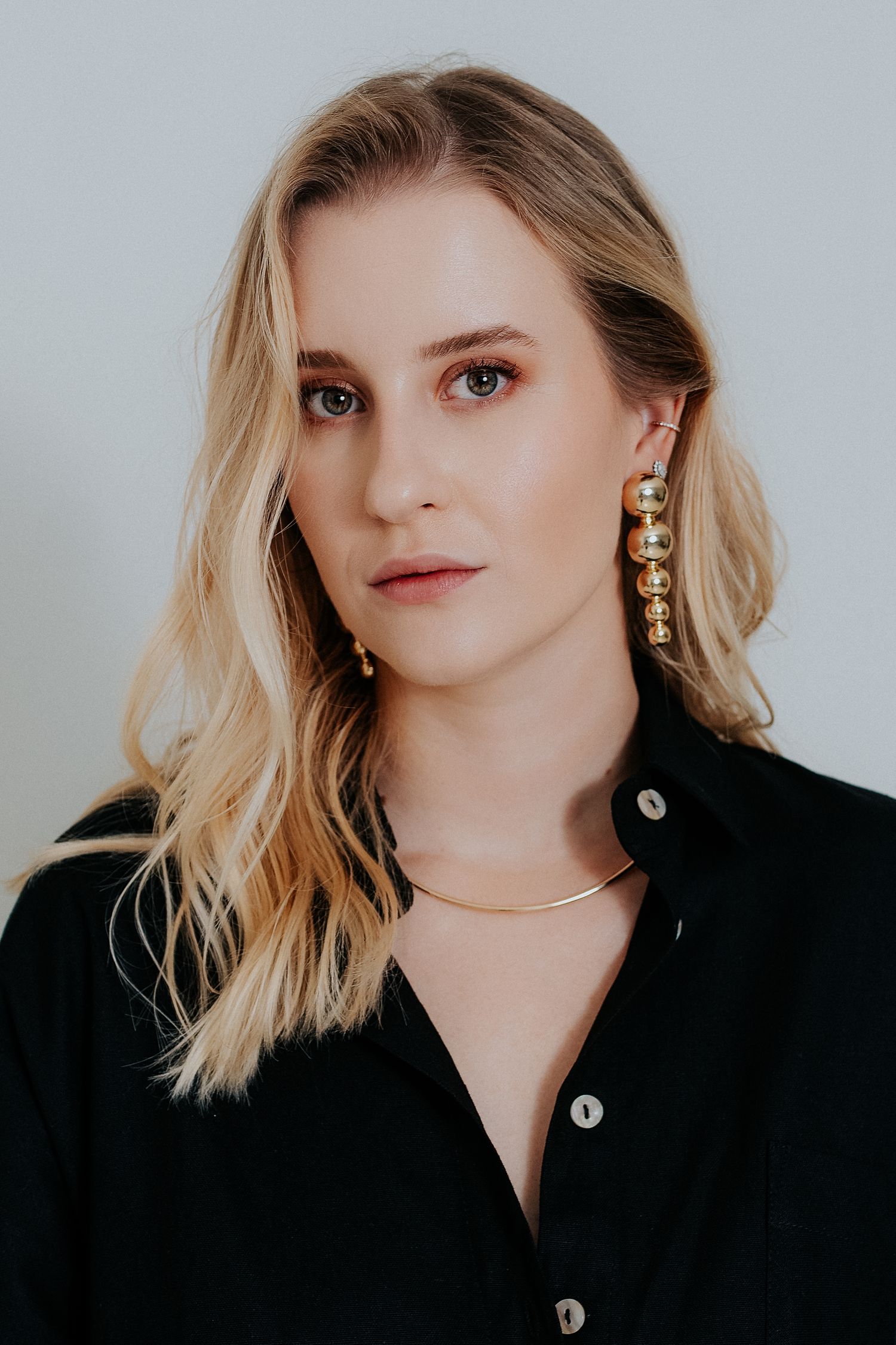 Portrait against light background wearing black button-up shirt and gold statement earrings with blonde hair.