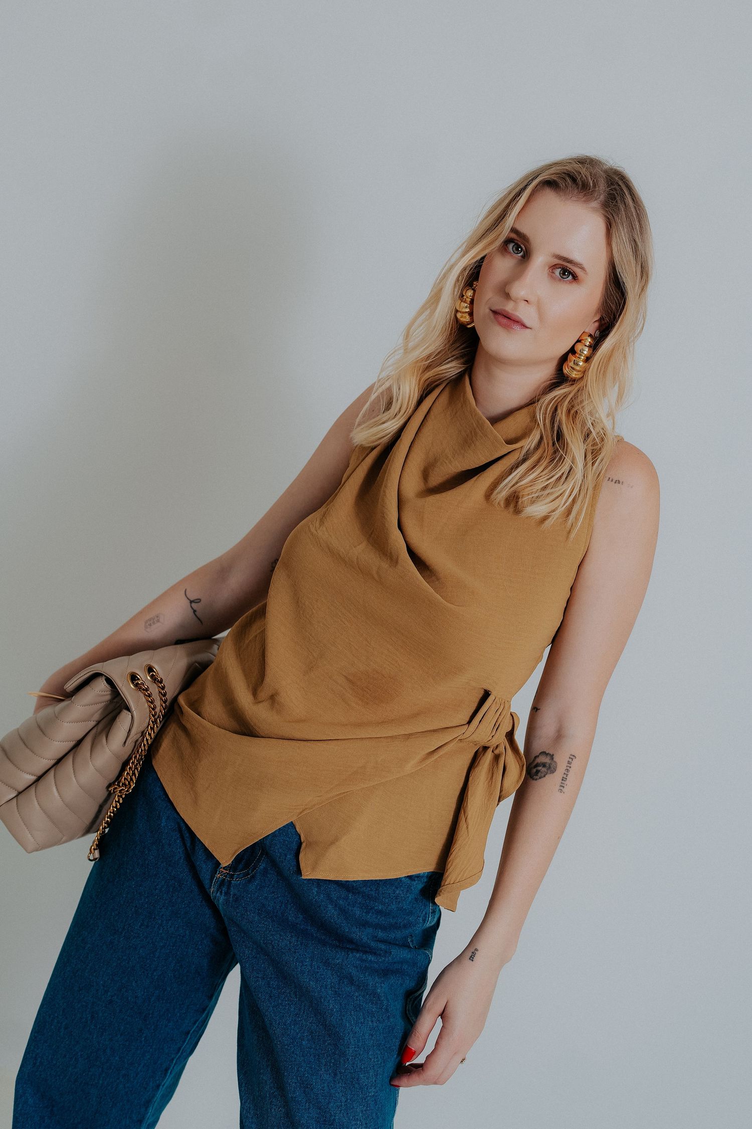 Woman in camel-colored sleeveless cowl neck top and jeans poses against light background.