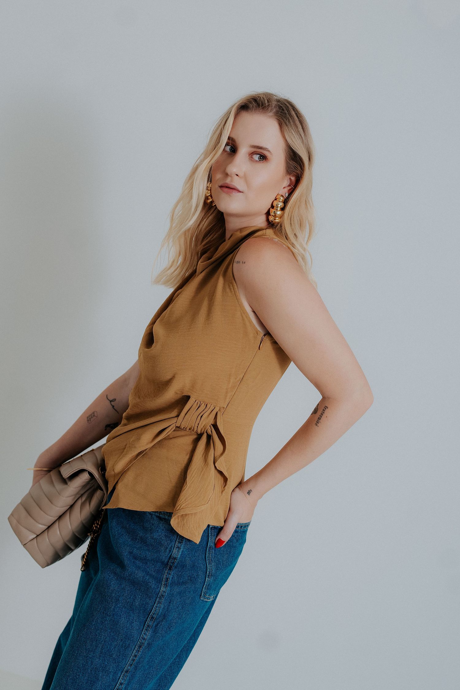 Model in a camel-colored sleeveless top and blue denim jeans poses in a minimalist setting.