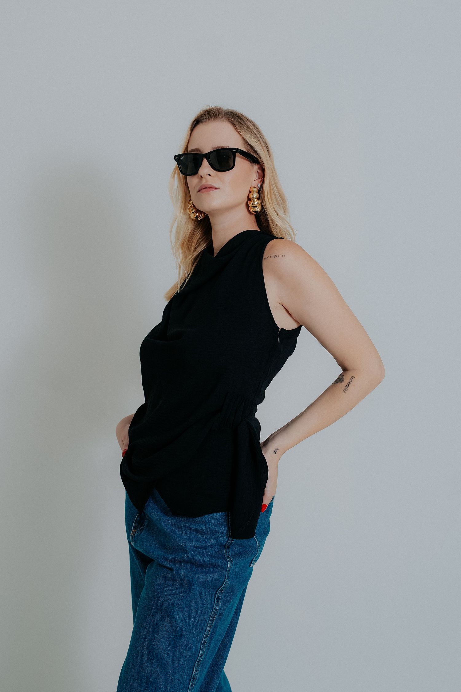 Stylish person in black sleeveless top and blue jeans poses confidently against white wall wearing dark sunglasses.