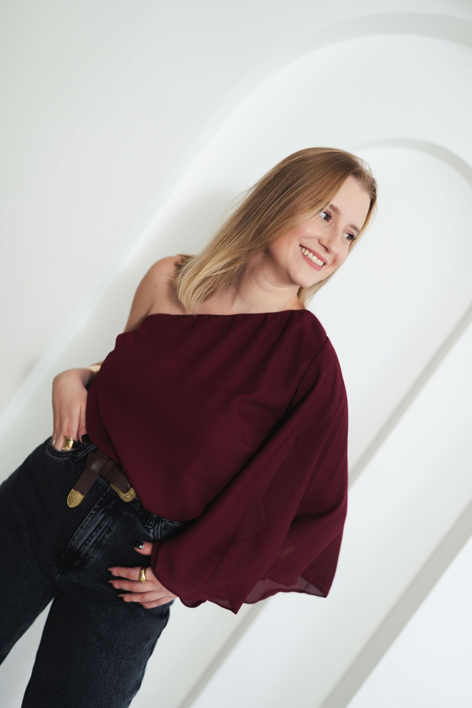 Model wearing burgundy off-shoulder sweater with dark jeans poses cheerfully against white background.