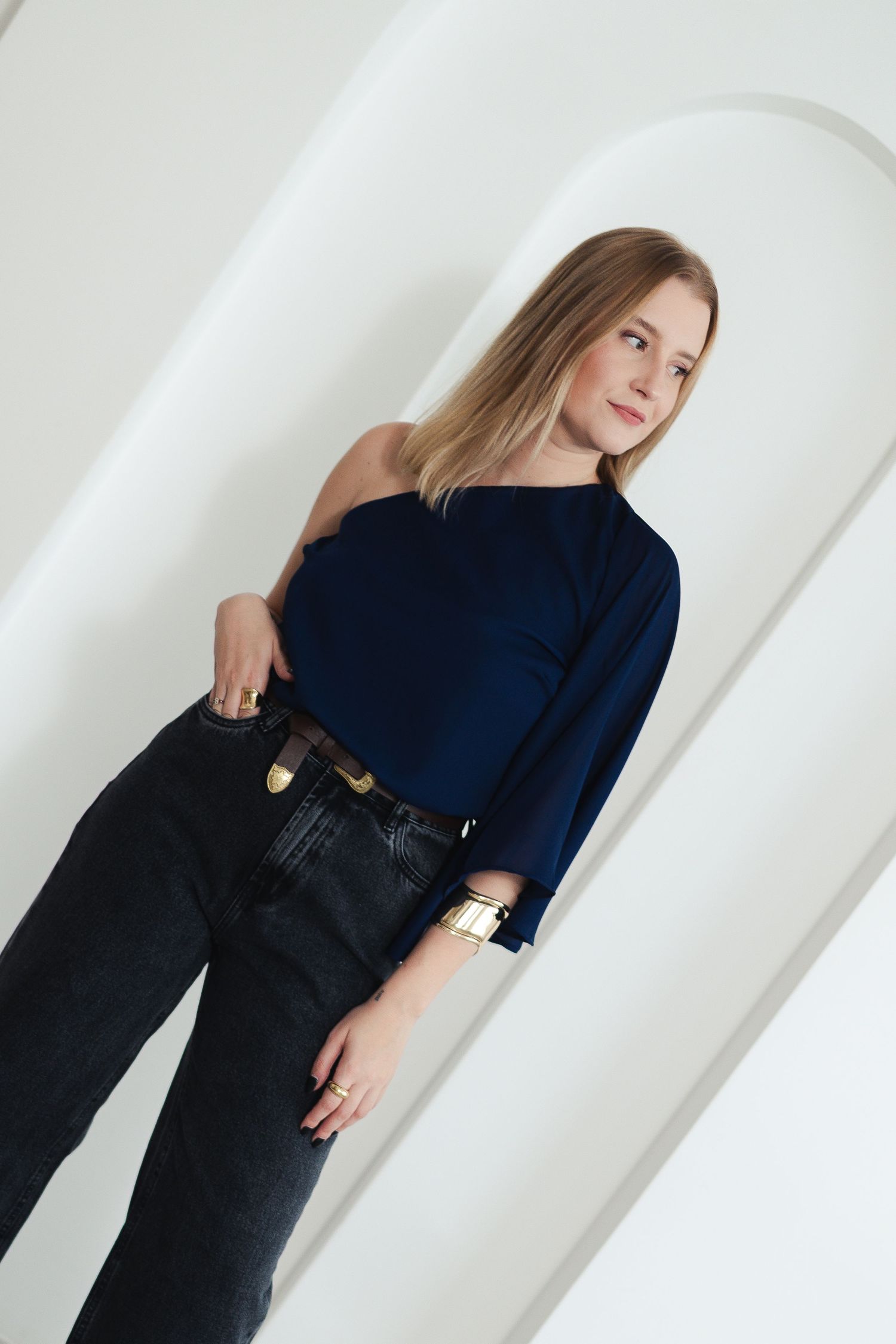 Model in navy blue top and dark denim poses elegantly against white arched wall.