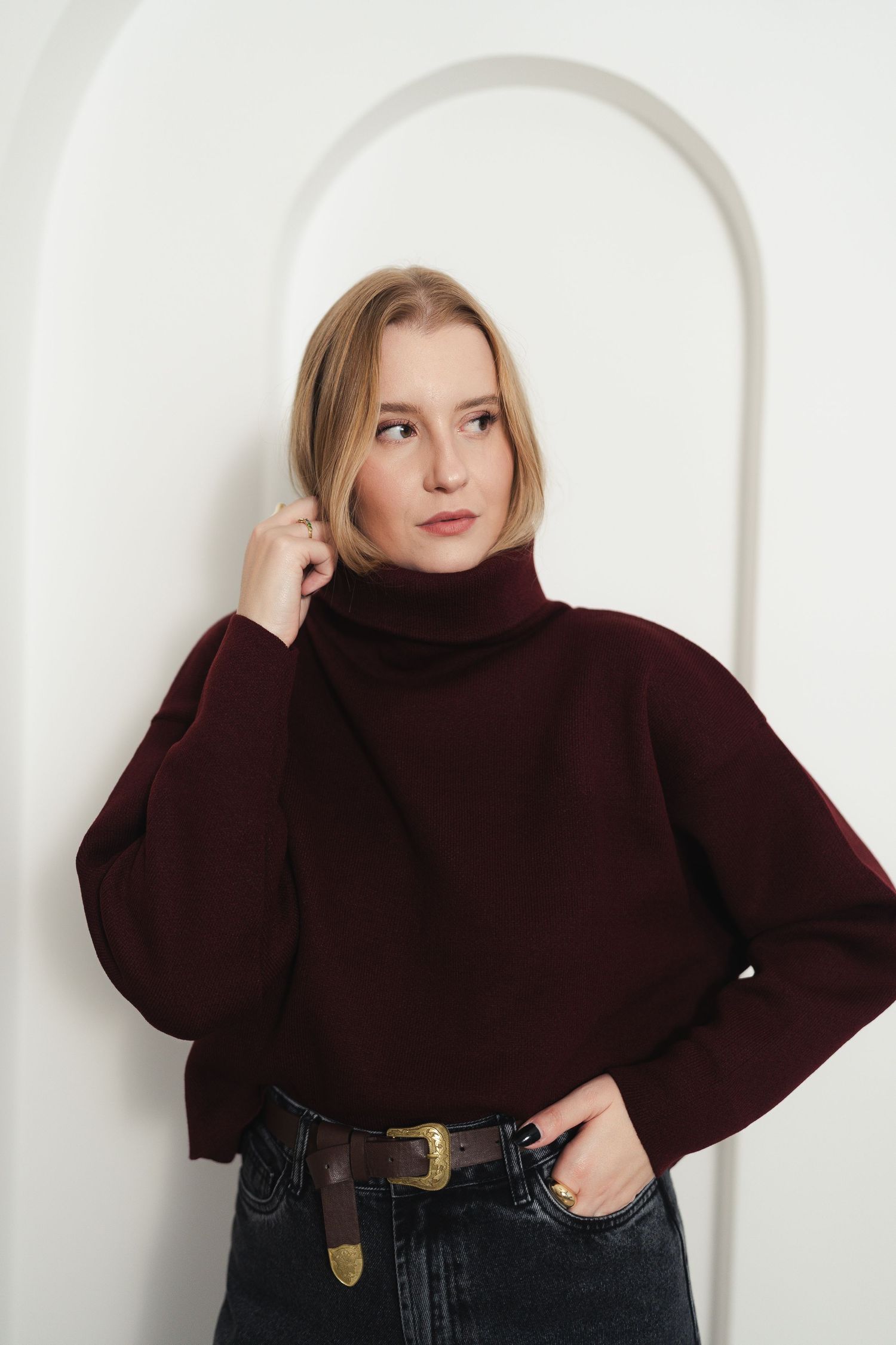 Fashion portrait of model in burgundy turtleneck sweater with belt detail against white arch.