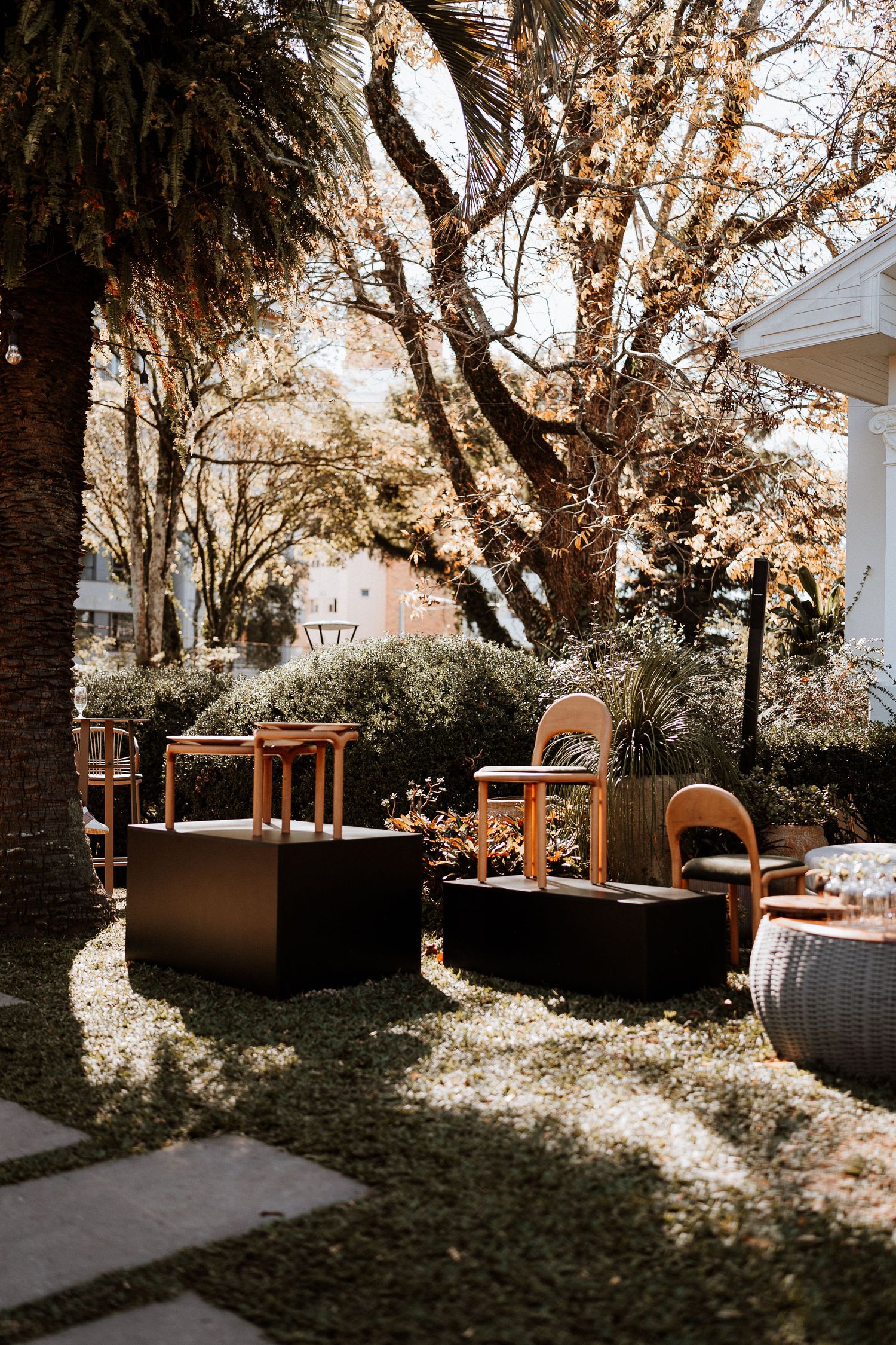 Outdoor seating area with wooden chairs and planters arranged along a white house exterior in dappled sunlight.