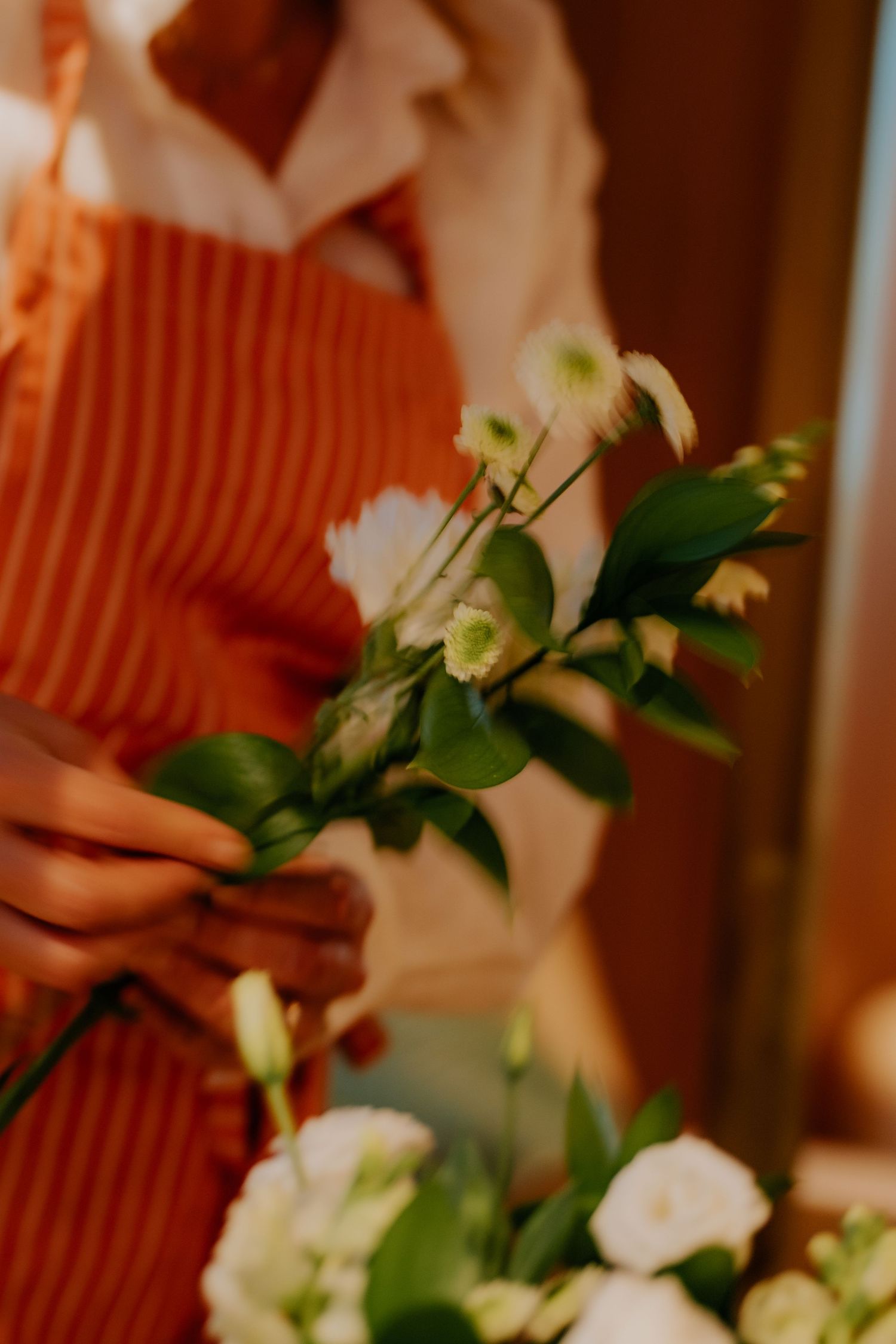 Hands in an orange sweater arrange delicate white flowers and greenery in a soft, warm-lit setting.