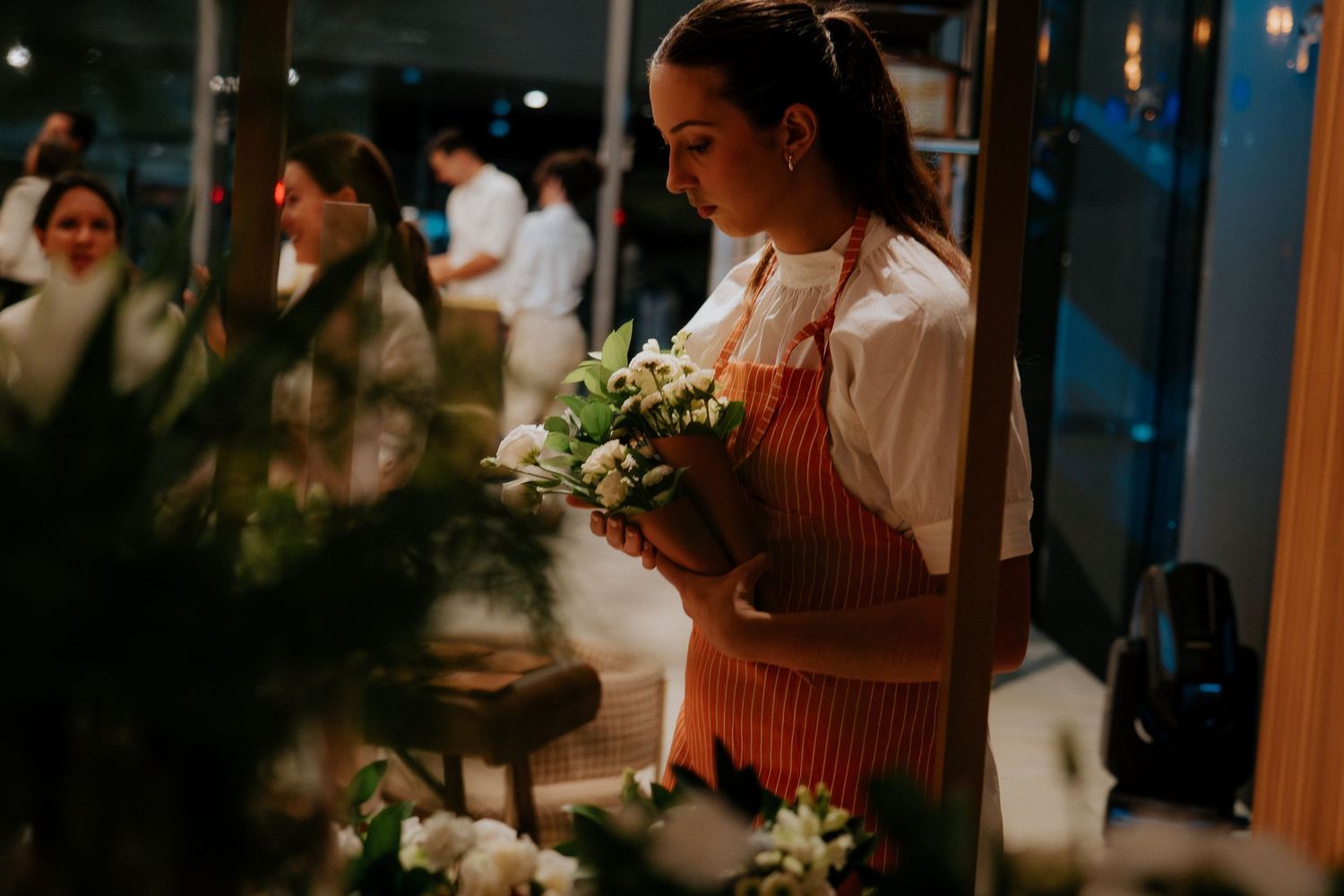 A florist in a red striped apron arranges white flowers in a dimly lit workshop space.