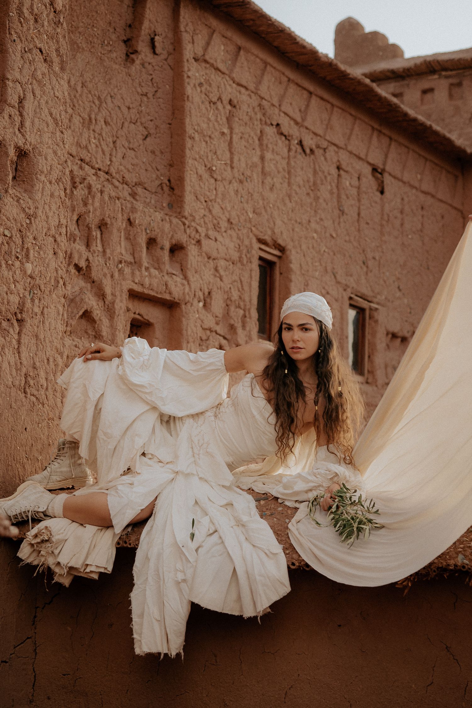 Woman in flowing white dress and beanie poses against adobe wall with fabric billowing in desert setting.